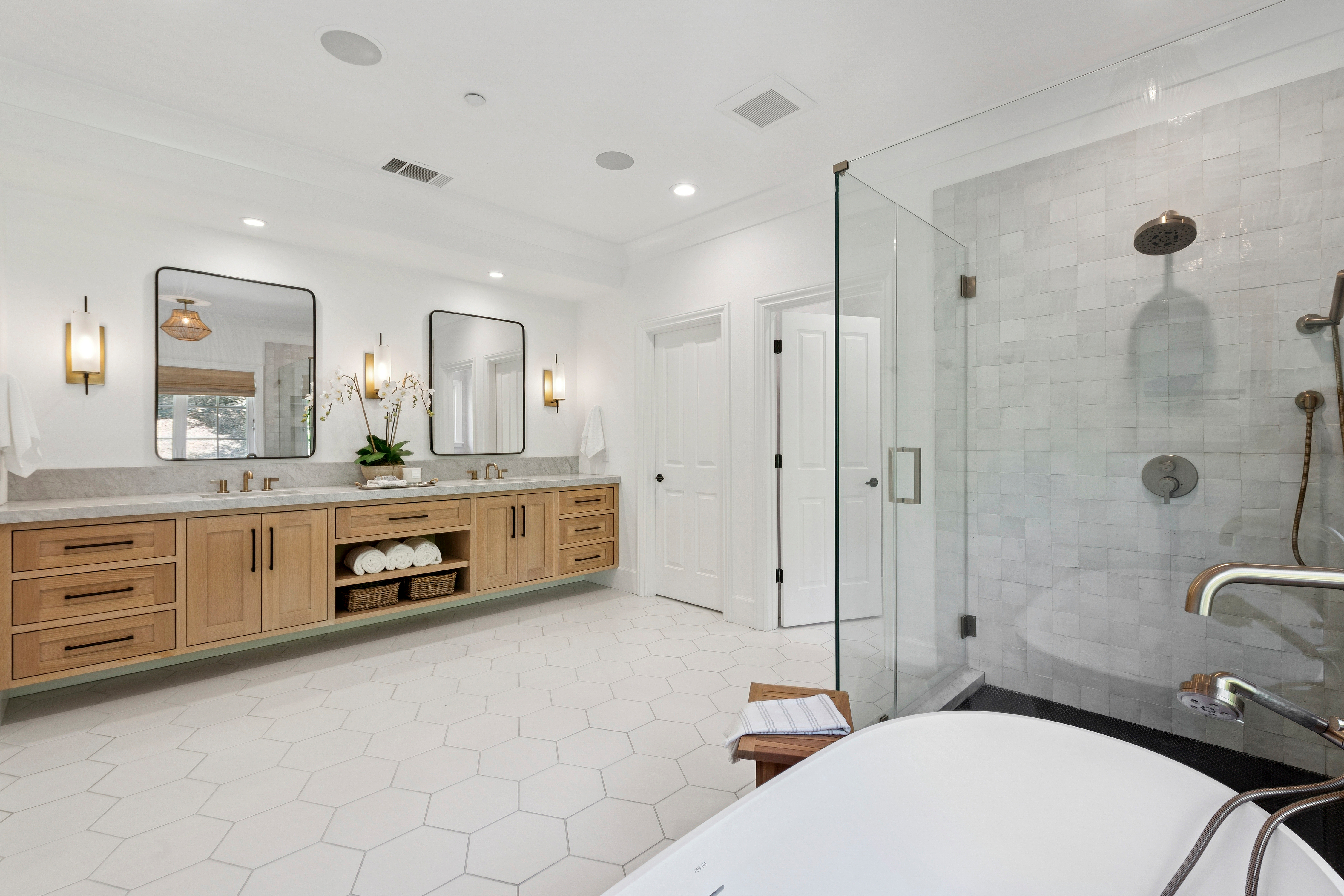 This spacious primary bathroom features a long, light wood vanity with dual sinks, topped with stone counters and framed by modern rectangular mirrors. To the right, a glass-enclosed shower with textured wall tiles sits adjacent to a freestanding soaking tub. The room is finished with large-format hexagonal floor tiles, creating a bright, contemporary, and spa-like atmosphere.