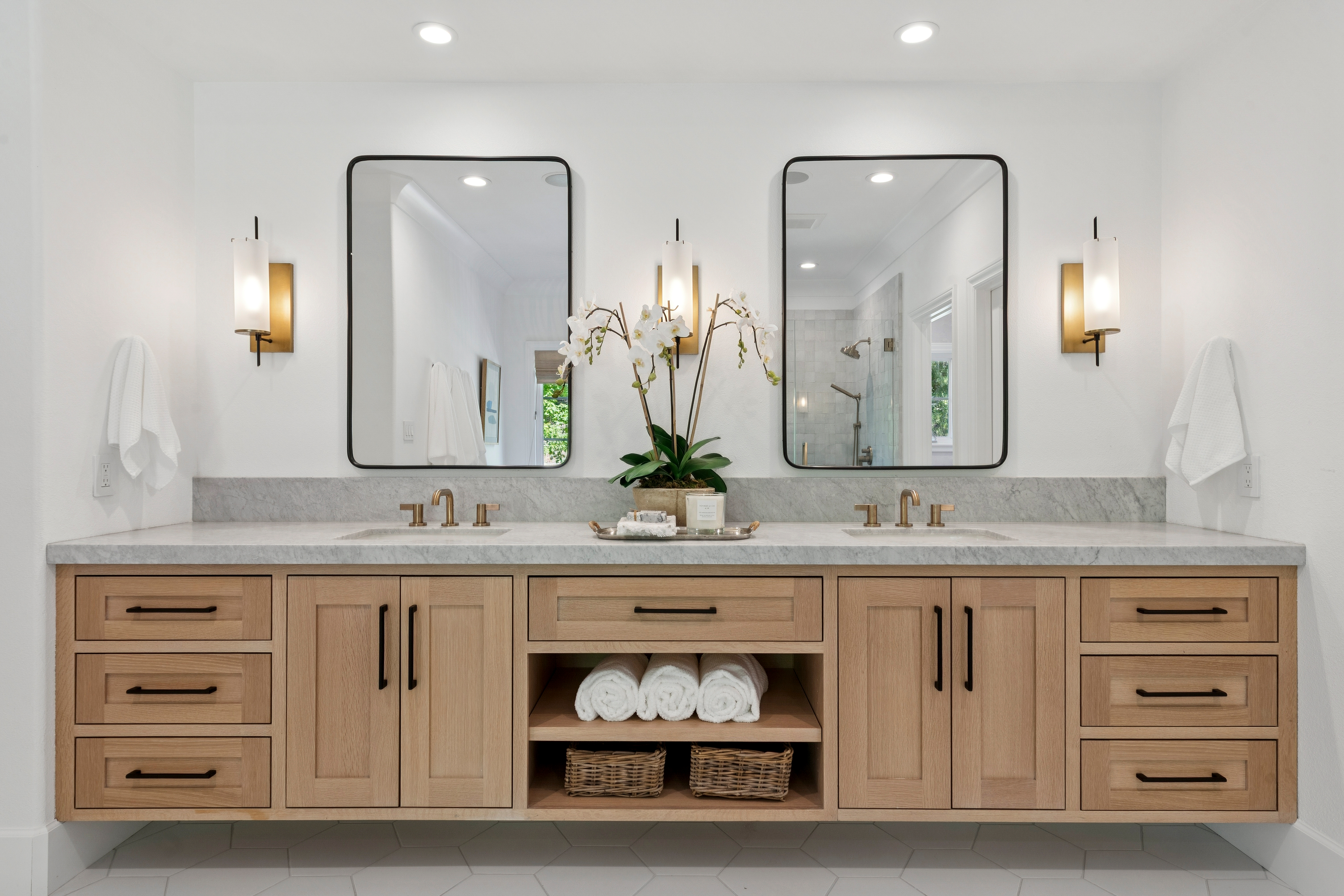This bright and airy primary bathroom features a double vanity with light wood cabinetry, topped with a neutral stone countertop and dual undermount sinks. Two rectangular mirrors with dark frames are mounted above the vanity, flanked by modern gold-toned wall sconces, while a central open shelving unit provides organized storage for towels and baskets. The space is accented by a potted orchid, creating a clean, sophisticated, and spa-like atmosphere.