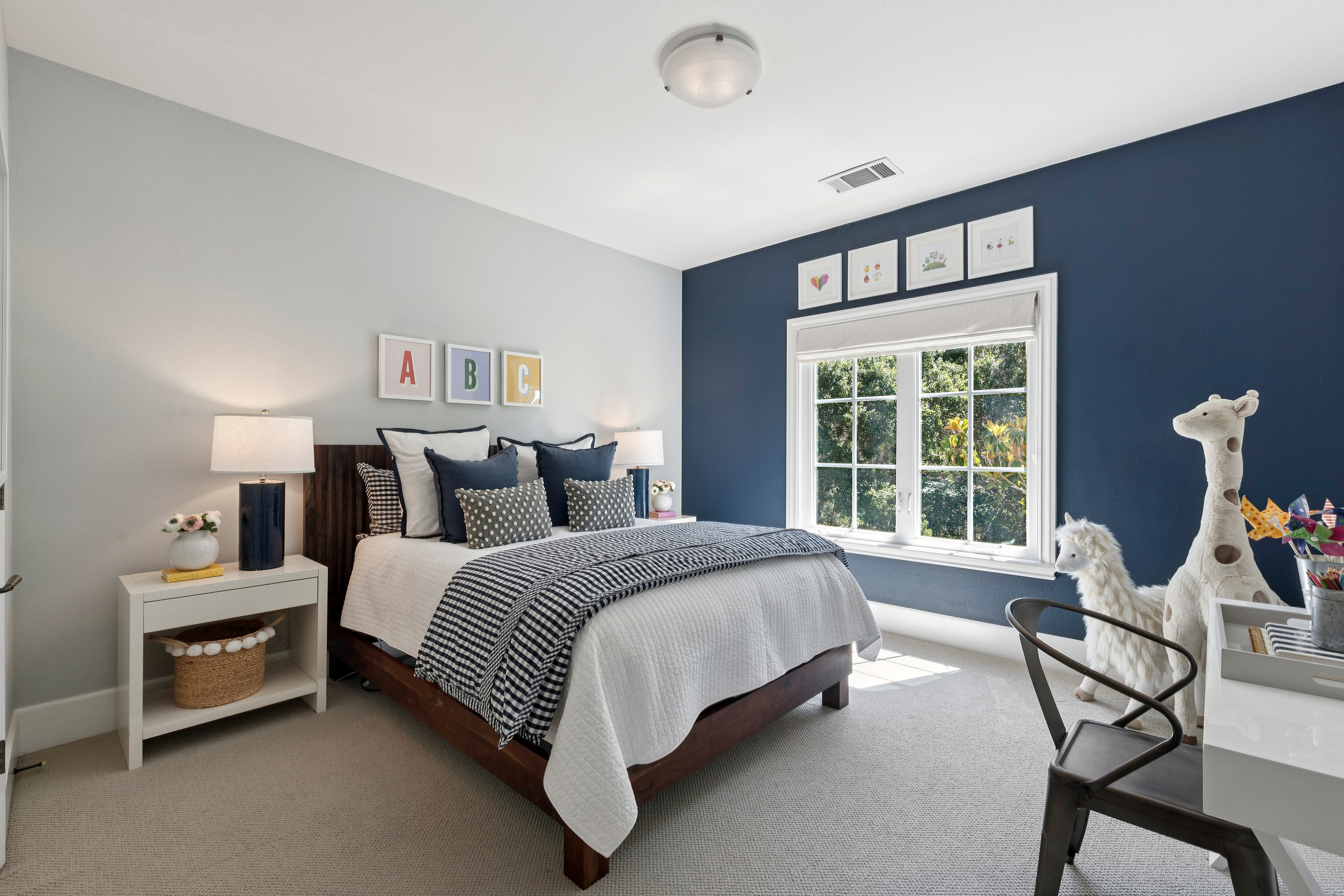 This charming child's bedroom features a bold navy accent wall contrasted by light gray walls and neutral carpeting. The room is furnished with a dark wood bed frame, a white nightstand, and a desk area, complemented by playful decor including framed alphabet art and stuffed animal figures. A large window with white trim allows natural light to brighten the space, creating a cozy and inviting atmosphere.