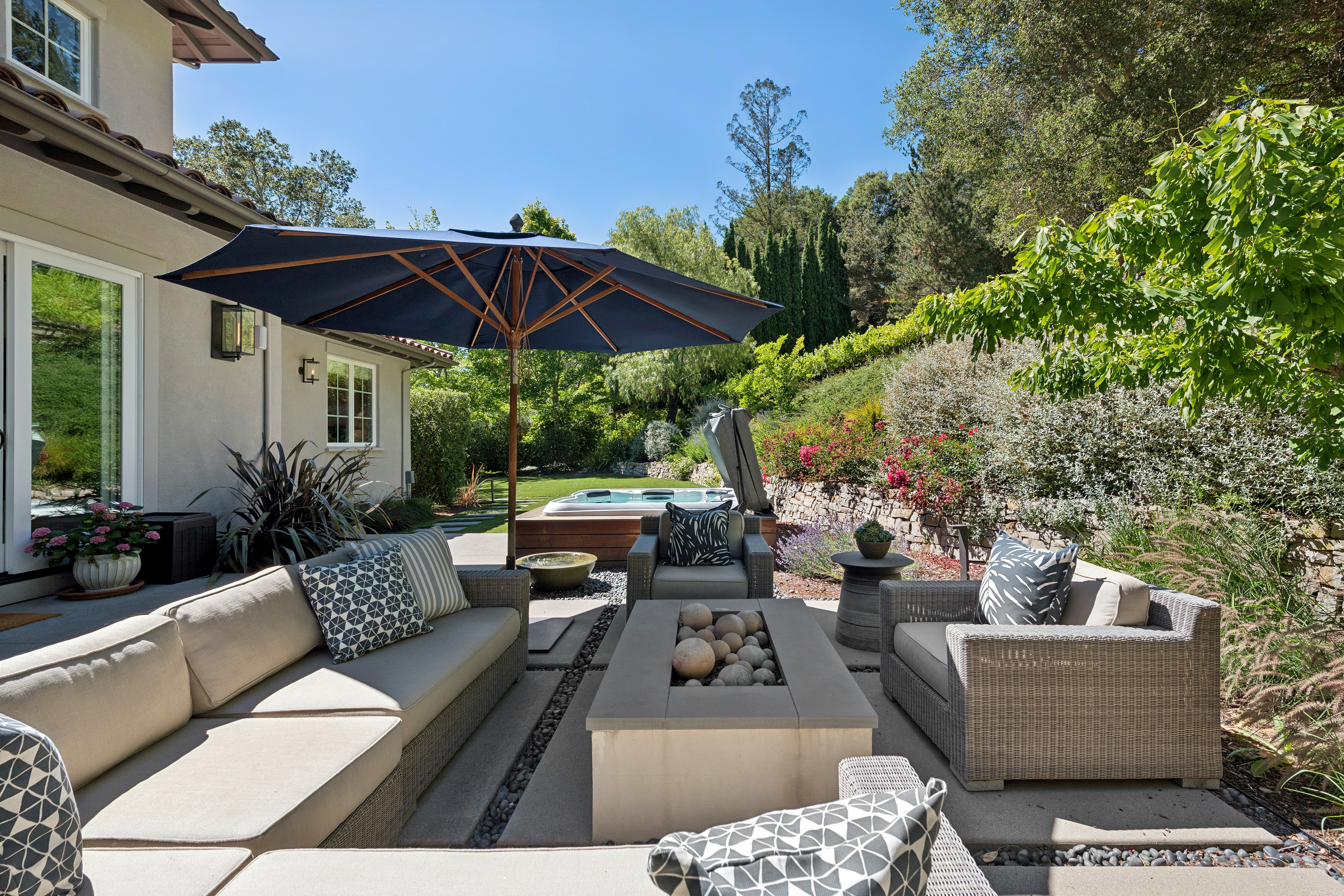 This inviting outdoor living space features a comfortable sectional sofa and two armchairs arranged around a rectangular fire pit filled with decorative stones. A large blue umbrella provides shade over the seating area, which is set on a concrete patio surrounded by a landscaped garden and a stone retaining wall. In the background, a private hot tub is nestled near the lush greenery, creating a serene and relaxing atmosphere perfect for entertaining.