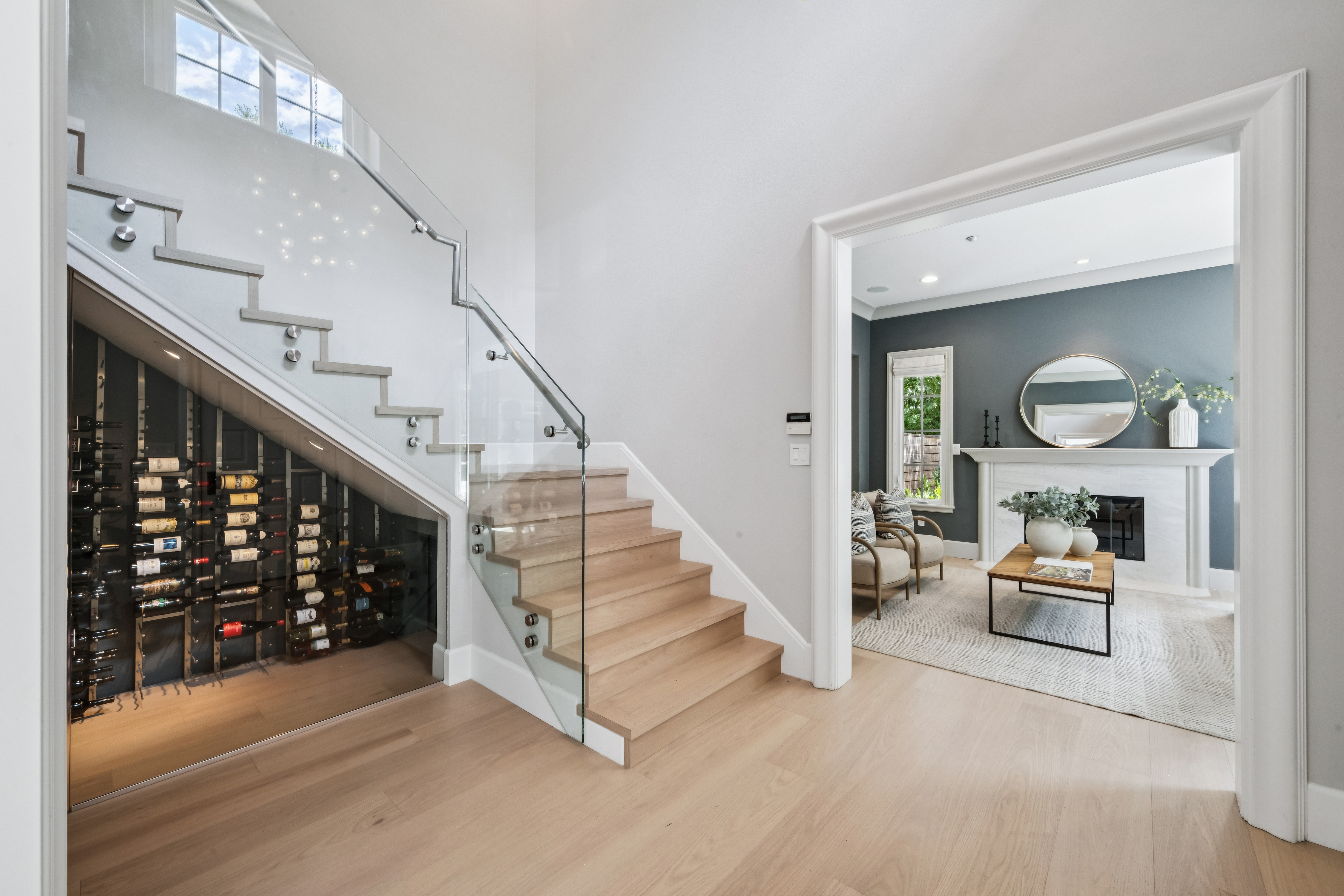 This bright entryway features a modern staircase with light wood treads and a sleek glass railing. Tucked neatly beneath the stairs is a custom-built wine cellar with metal racking, while an adjacent doorway leads into a cozy sitting room with a fireplace and dark accent walls. The open floor plan and neutral color palette create a sophisticated and inviting atmosphere.