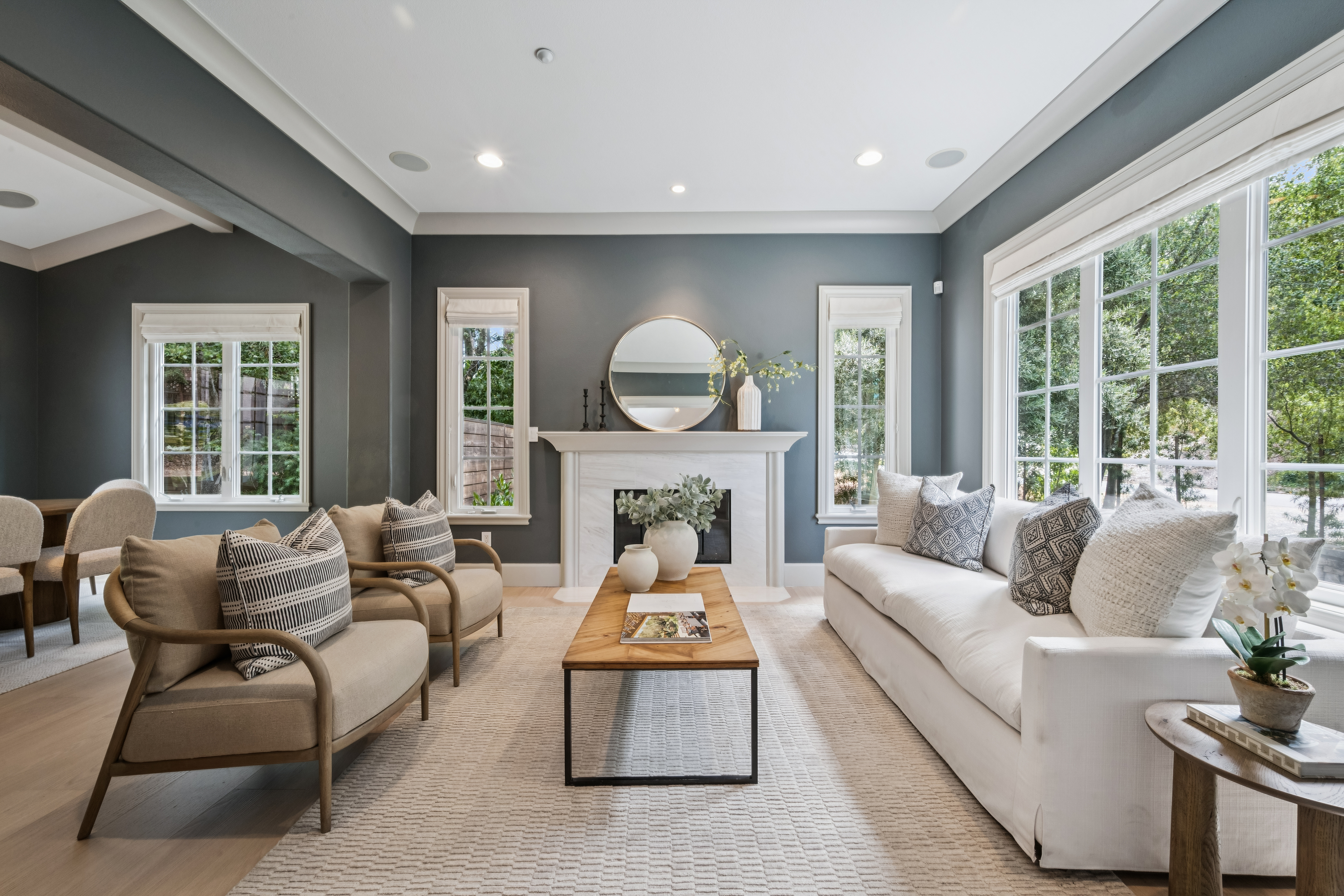 This bright and inviting living room features deep blue walls contrasted by a white fireplace mantel and large windows that flood the space with natural light. The room is furnished with a plush white sofa, two light-toned armchairs with patterned throw pillows, and a central wood-topped coffee table resting on a textured area rug. The overall aesthetic is sophisticated and airy, creating a comfortable atmosphere perfect for relaxation or entertaining.