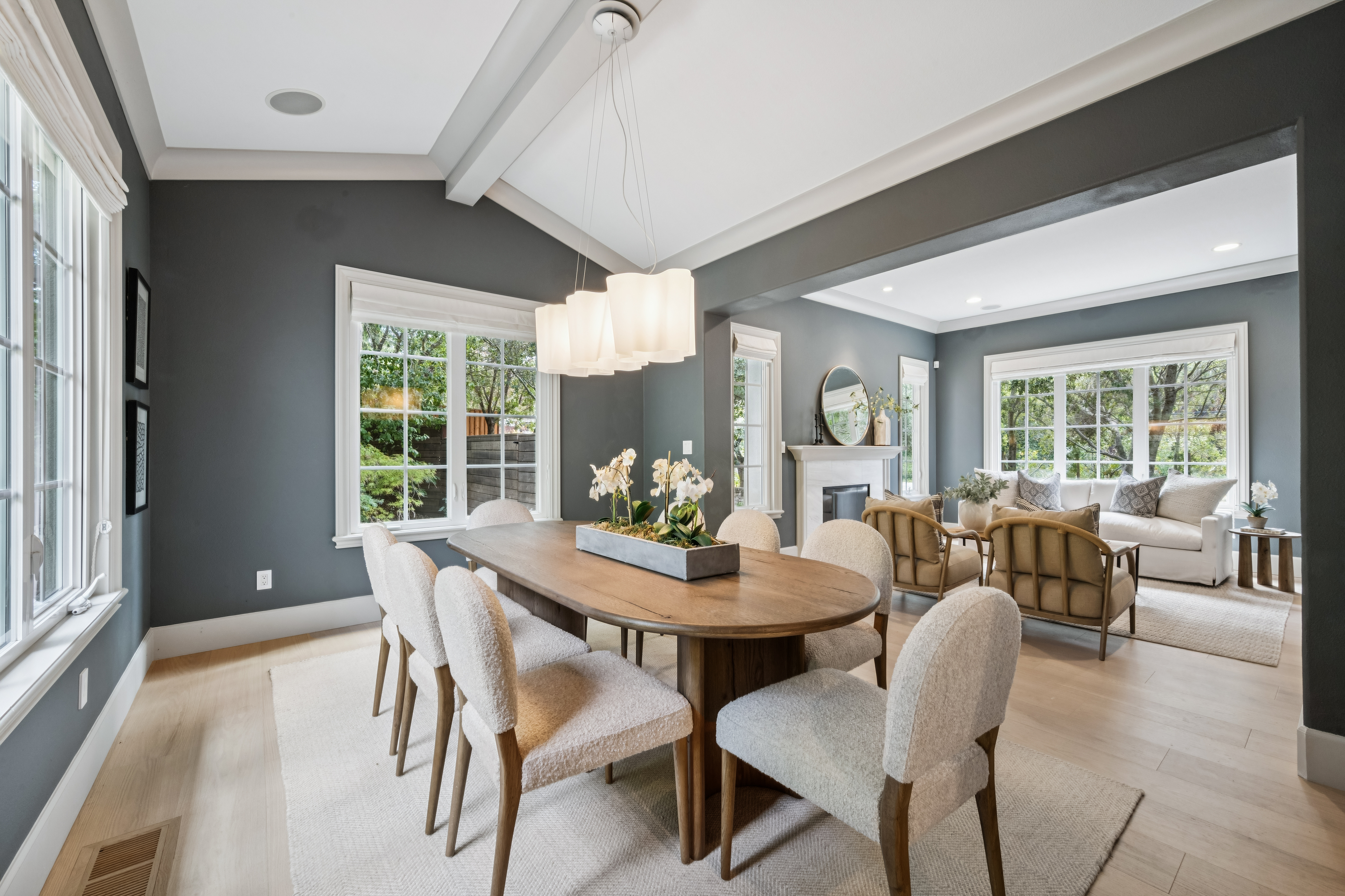 This elegant dining room features a large oval wooden table surrounded by six upholstered chairs, set beneath a modern multi-pendant light fixture. The space is painted in a sophisticated dark gray, which contrasts beautifully with the light wood flooring and white trim. An open floor plan allows for a seamless transition into the adjacent living area, where a fireplace with a white mantel serves as a focal point.