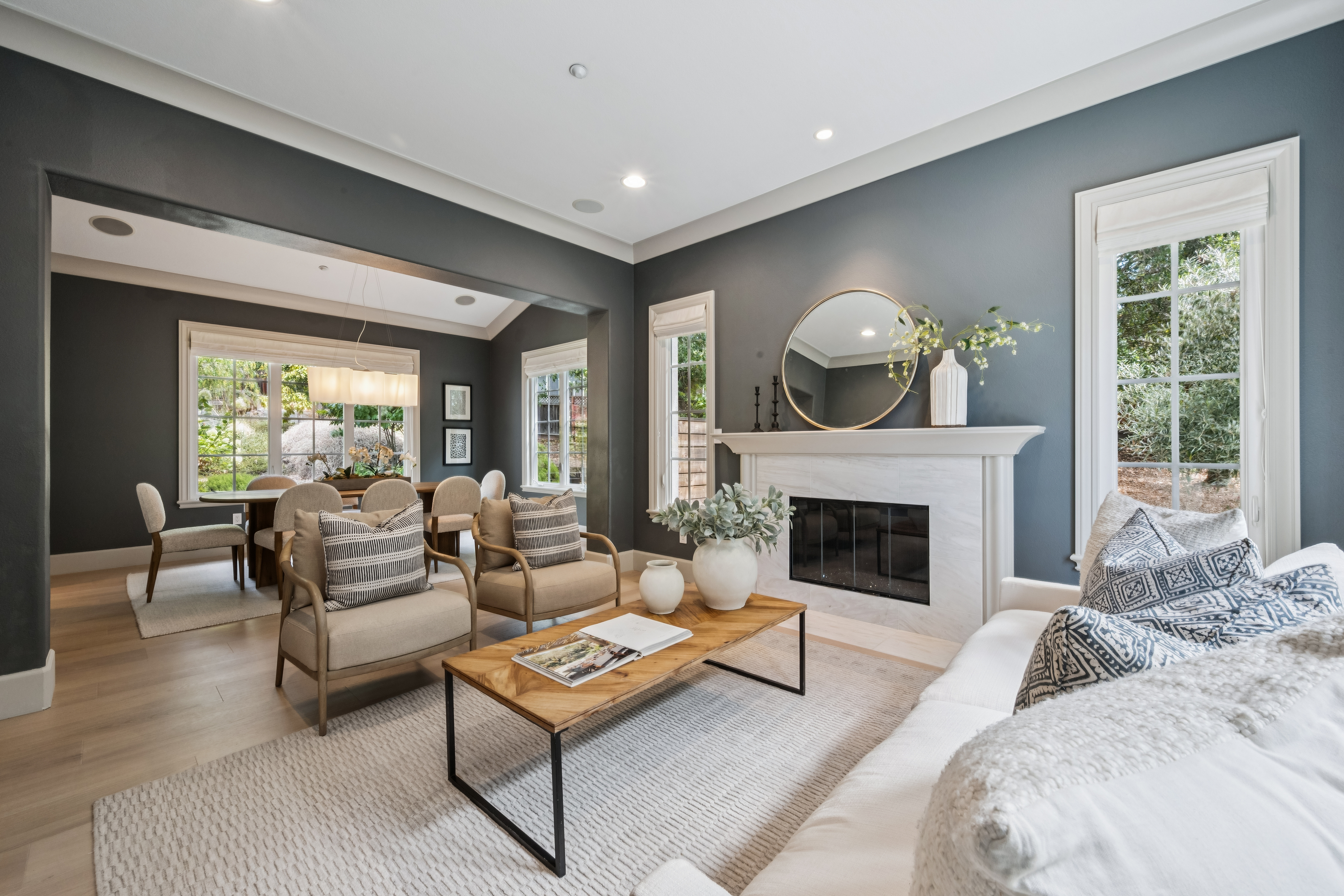 This bright and inviting living room features a neutral color palette accented by deep blue-grey walls and light wood flooring. A central fireplace with a stone surround and a round mirror serves as the focal point, while a comfortable seating area with a wooden coffee table and patterned throw pillows creates a cozy atmosphere. The space flows seamlessly into an adjacent dining area, with large windows providing ample natural light and views of the surrounding greenery.