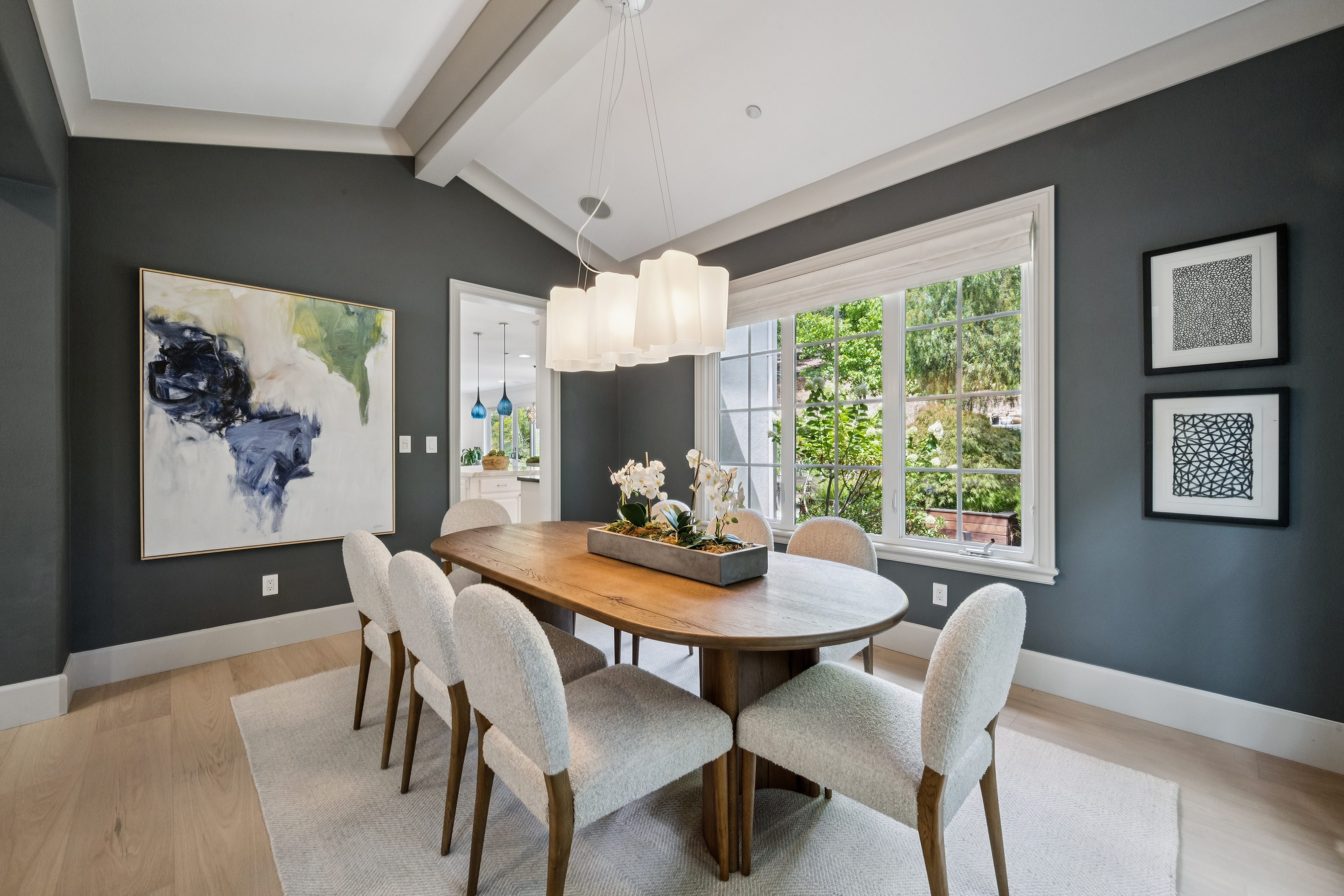 This elegant dining room features dark charcoal walls contrasted by light wood flooring and a large, oval wooden dining table surrounded by six textured, cream-colored chairs. A modern, multi-tiered pendant light fixture hangs above a central floral centerpiece, while large windows provide ample natural light and a view of the greenery outside. The space is decorated with a large abstract painting on the left wall and two framed geometric art pieces on the right, creating a sophisticated and inviting atmosphere.