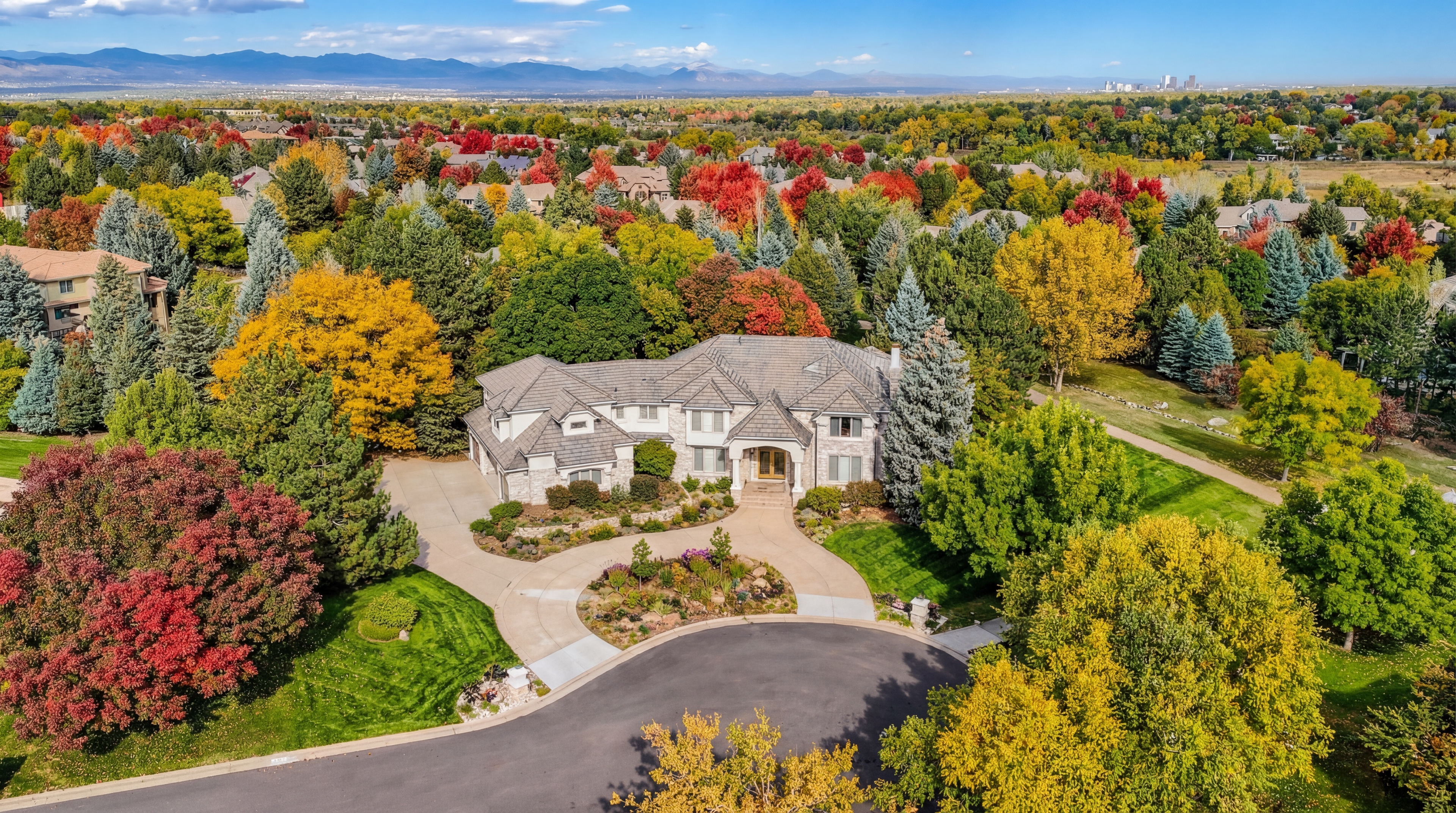 This stunning aerial perspective showcases a large, multi-gabled stone residence set within a lush, mature landscape vibrant with autumn foliage. The property features a spacious, landscaped circular driveway, manicured green lawns, and expansive views reaching back toward a city skyline and mountain range. The home exudes luxury and curb appeal, perfectly integrated into a wooded, serene residential neighborhood.