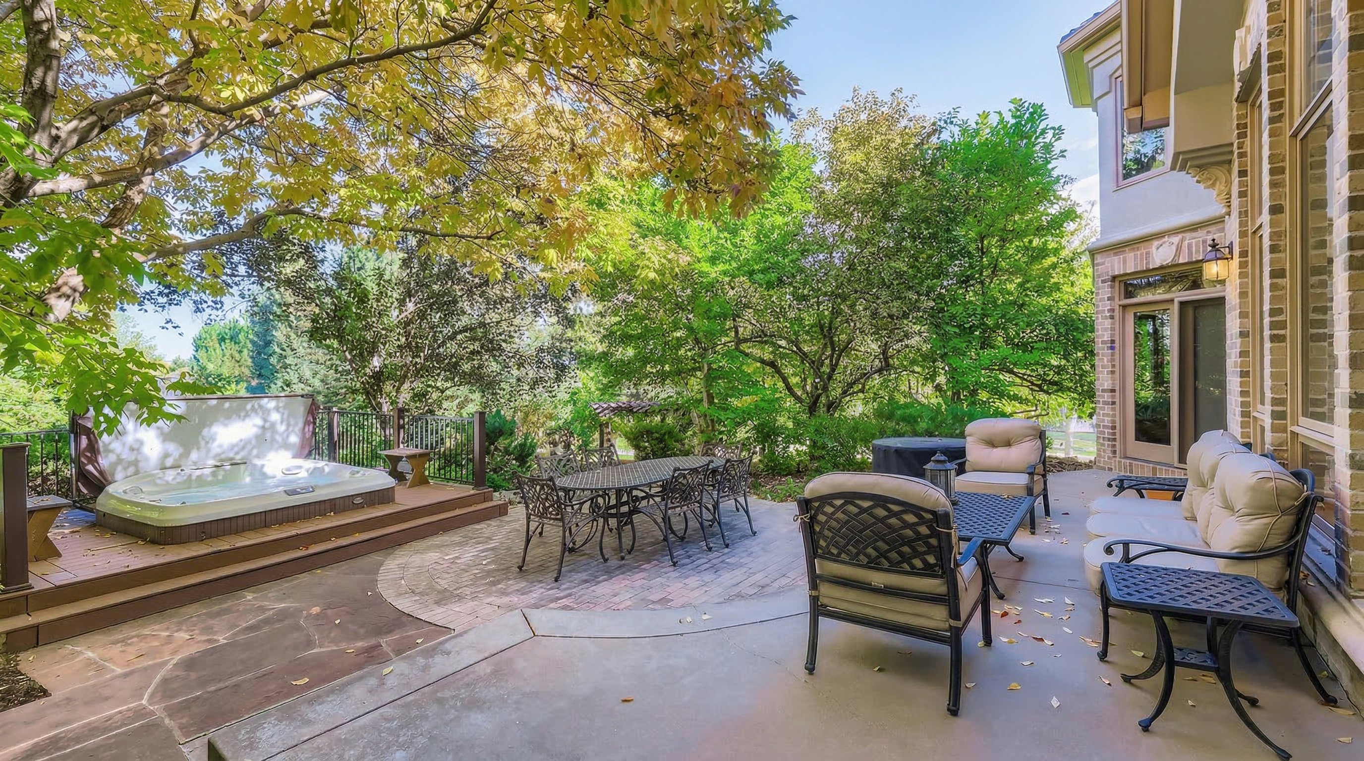 This spacious outdoor living area features a tiered design with a wooden deck housing a built-in hot tub alongside a large stone-paver patio. The space is furnished with an elegant wrought-iron dining set and comfortable cushioned seating, perfect for al fresco dining and relaxation. Surrounded by lush mature trees and greenery, the patio provides a private, inviting environment directly accessible from the main house.