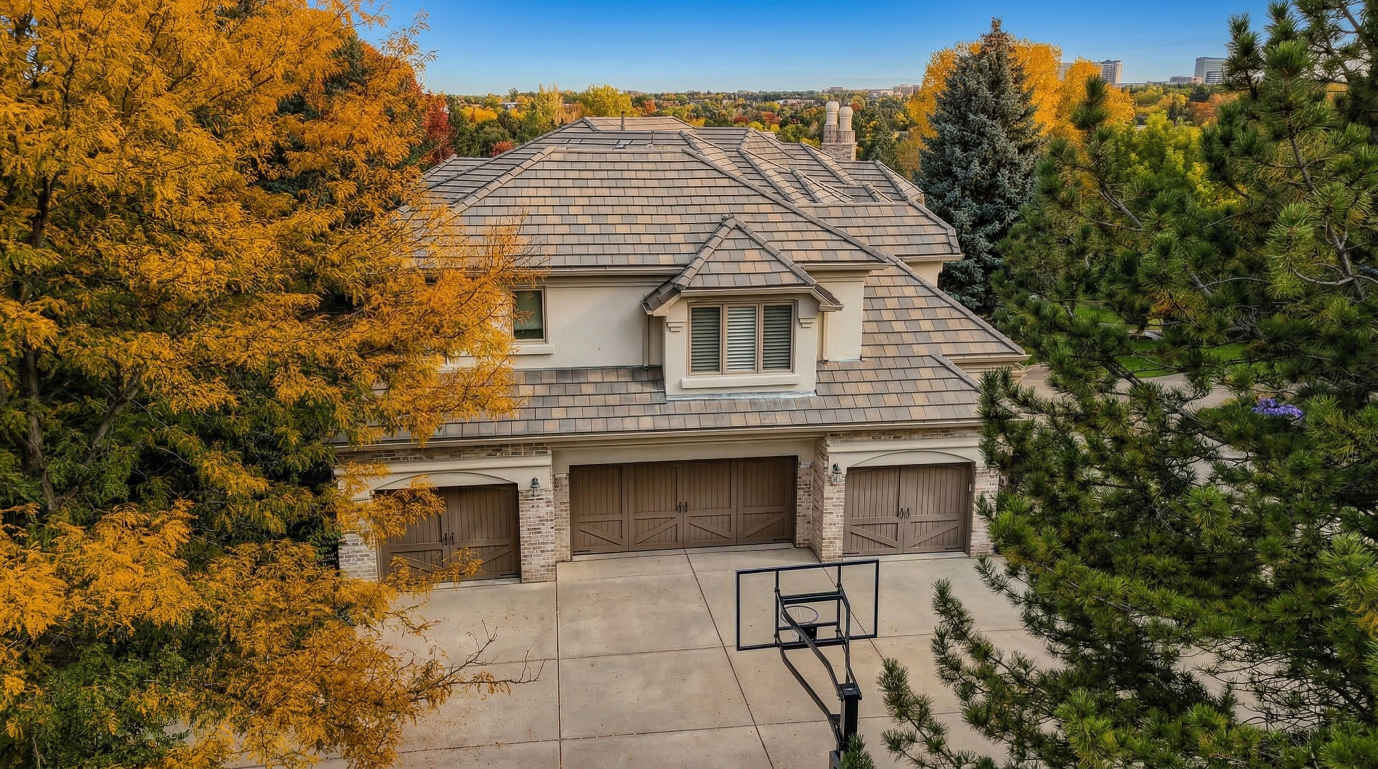 This aerial view showcases a stately, traditional home featuring a multi-car garage with custom wood carriage-style doors and a spacious concrete driveway. The exterior is accented with brick and stone finishes, complemented by a textured tile roof and mature, vibrant fall foliage. A basketball hoop is positioned on the driveway, adding a functional recreational element to this well-maintained suburban property.