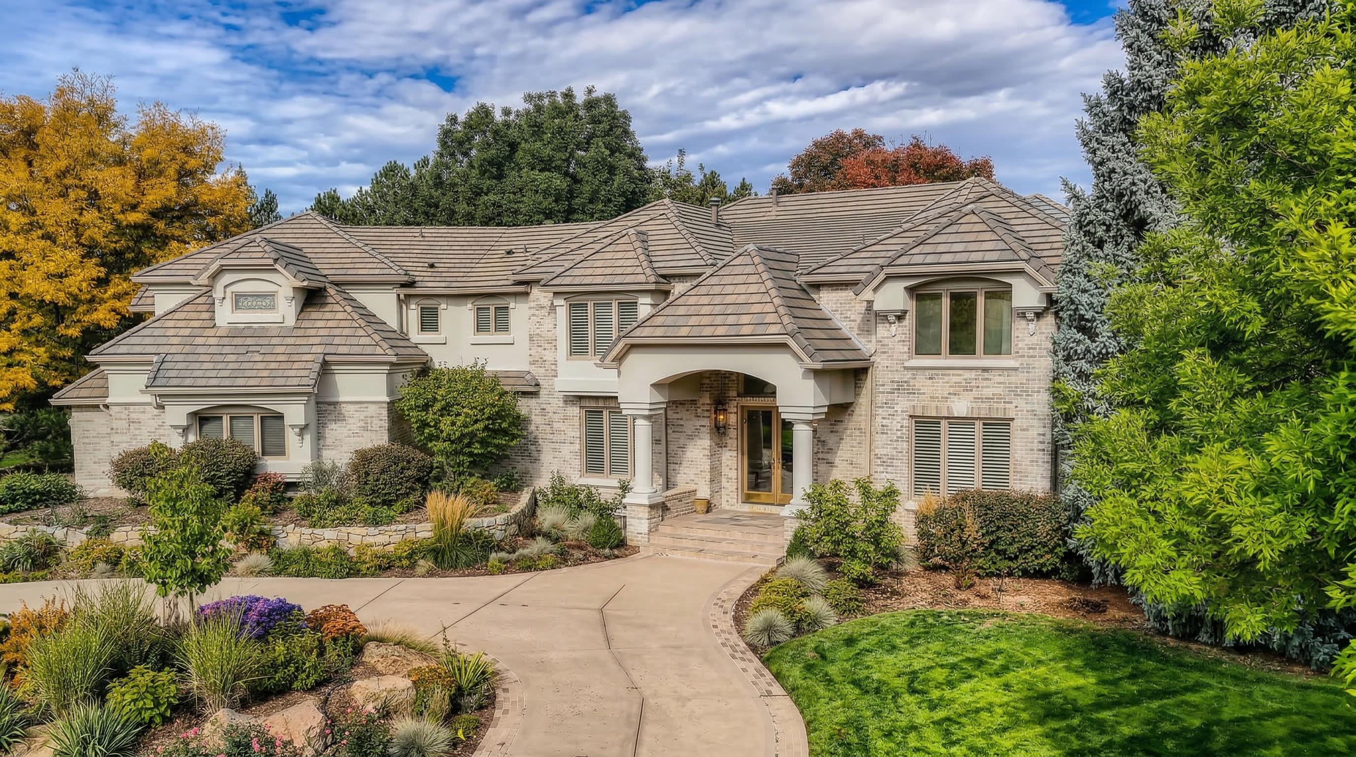 This grand, two-story residence features a sophisticated light brick exterior accented by multi-layered pitched rooflines and an elegant, columned front portico. Surrounded by lush, professional landscaping and mature trees, the home offers a welcoming and stately curb appeal enhanced by a curved driveway. The property balances architectural detail and natural beauty, presenting an upscale suburban aesthetic.