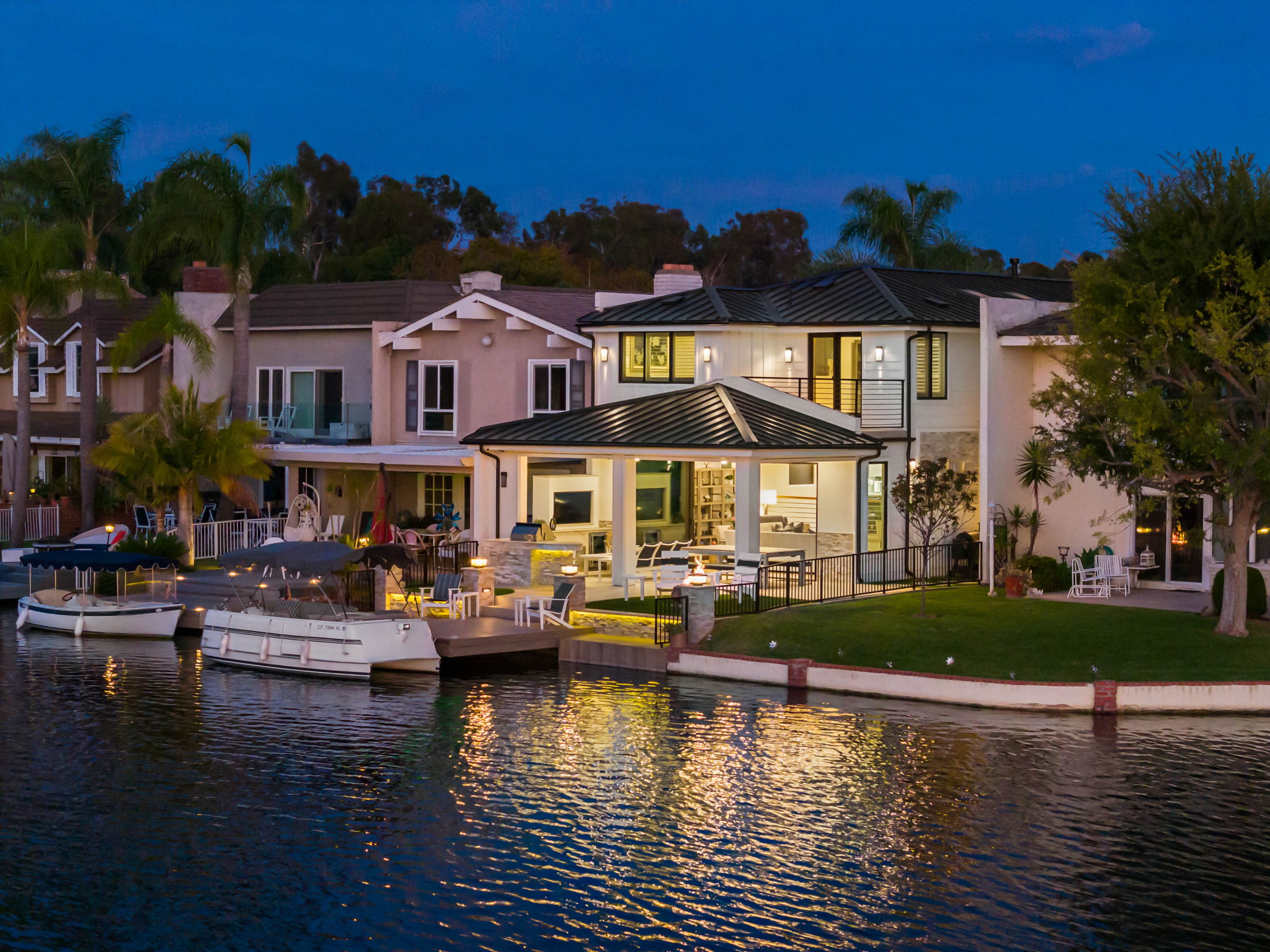 This stunning waterfront property features a beautifully designed modern home with a spacious covered outdoor patio and private boat dock. The residence highlights contemporary architecture with clean lines, soft evening lighting, and seamless indoor-outdoor transition, perfectly positioned on a calm canal. The lush lawn, mature landscaping, and reflective water create an inviting and tranquil atmosphere.