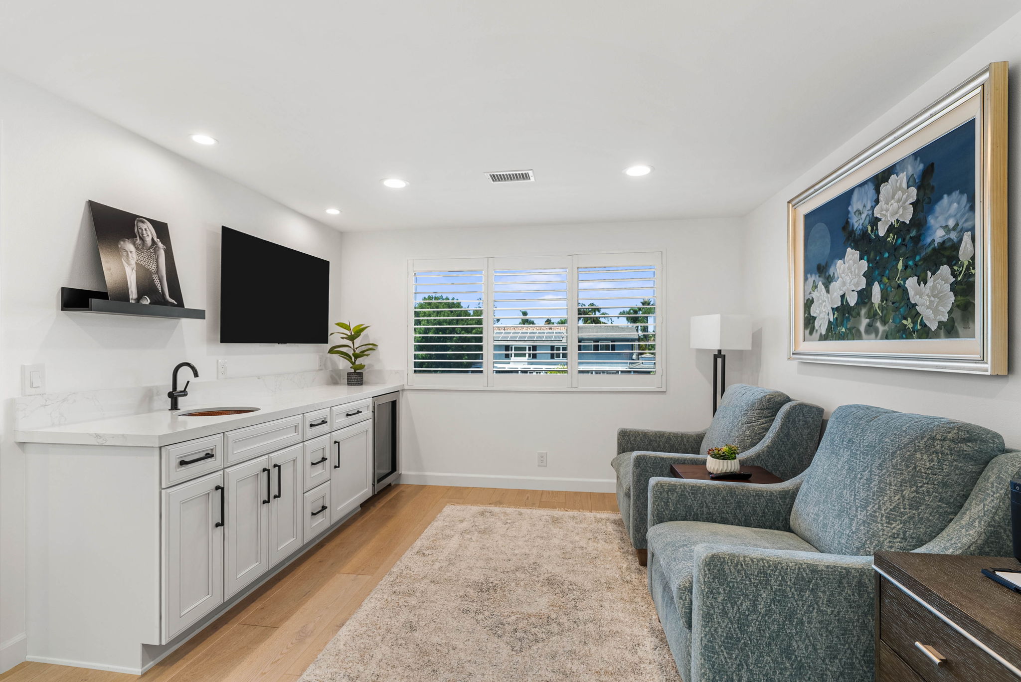 This cozy and functional living space features a wet bar area with white cabinetry, a marble countertop, and a built-in wine cooler, perfect for entertaining. The room is comfortably furnished with two blue upholstered armchairs, soft textured area rug, and a large decorative floral artwork on the wall. The space is well-lit with recessed lighting and benefits from natural light streaming through the central plantation-shuttered window.