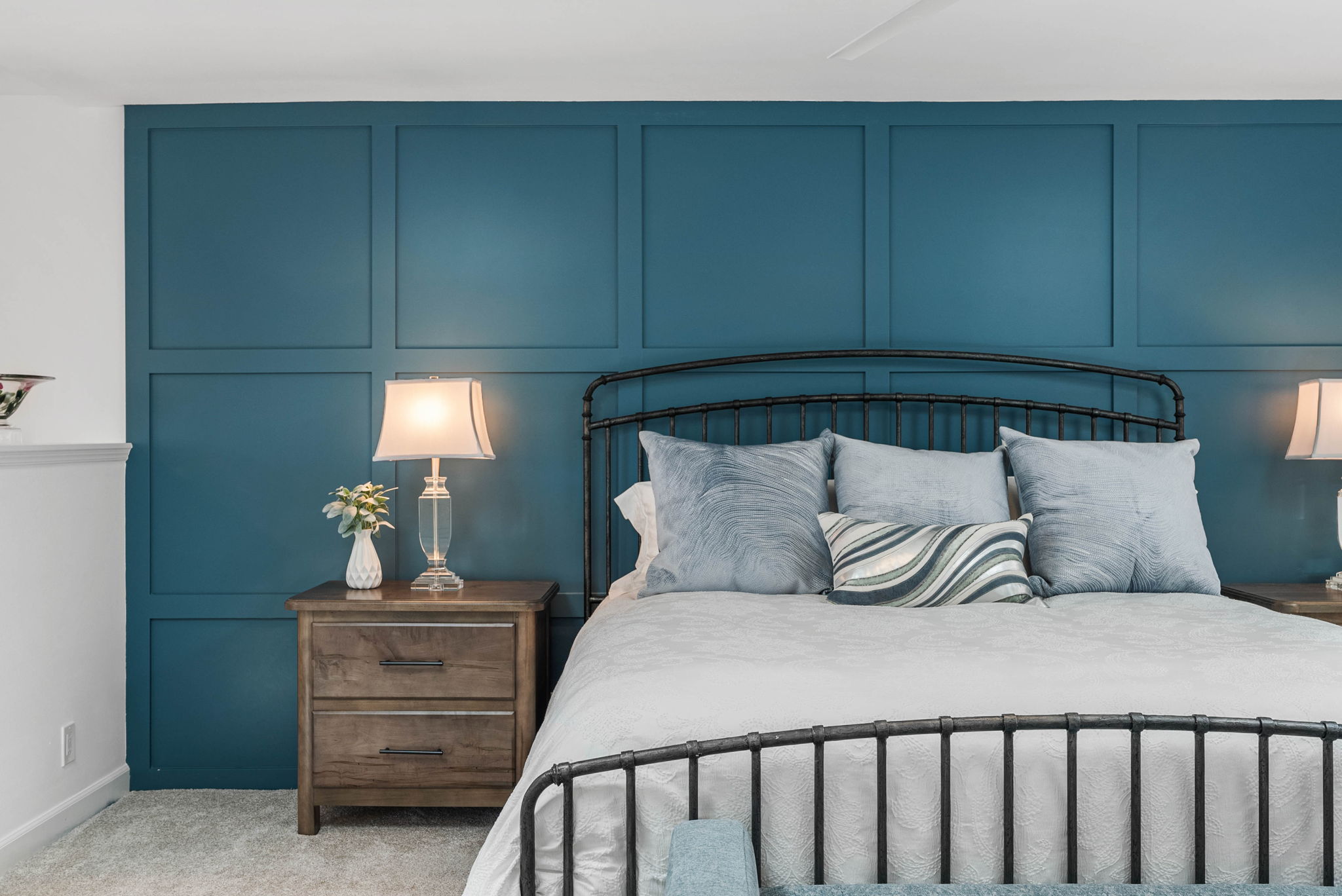 This primary bedroom features a striking deep teal accent wall with a sophisticated wainscoting grid pattern, serving as a focal point behind a dark metal bed frame. A wooden bedside table with a crystal-based lamp adds a touch of elegance, while plush pillows in shades of blue and gray enhance the room's serene, upscale atmosphere. The overall aesthetic combines modern classic design elements for a cozy and inviting retreat.
