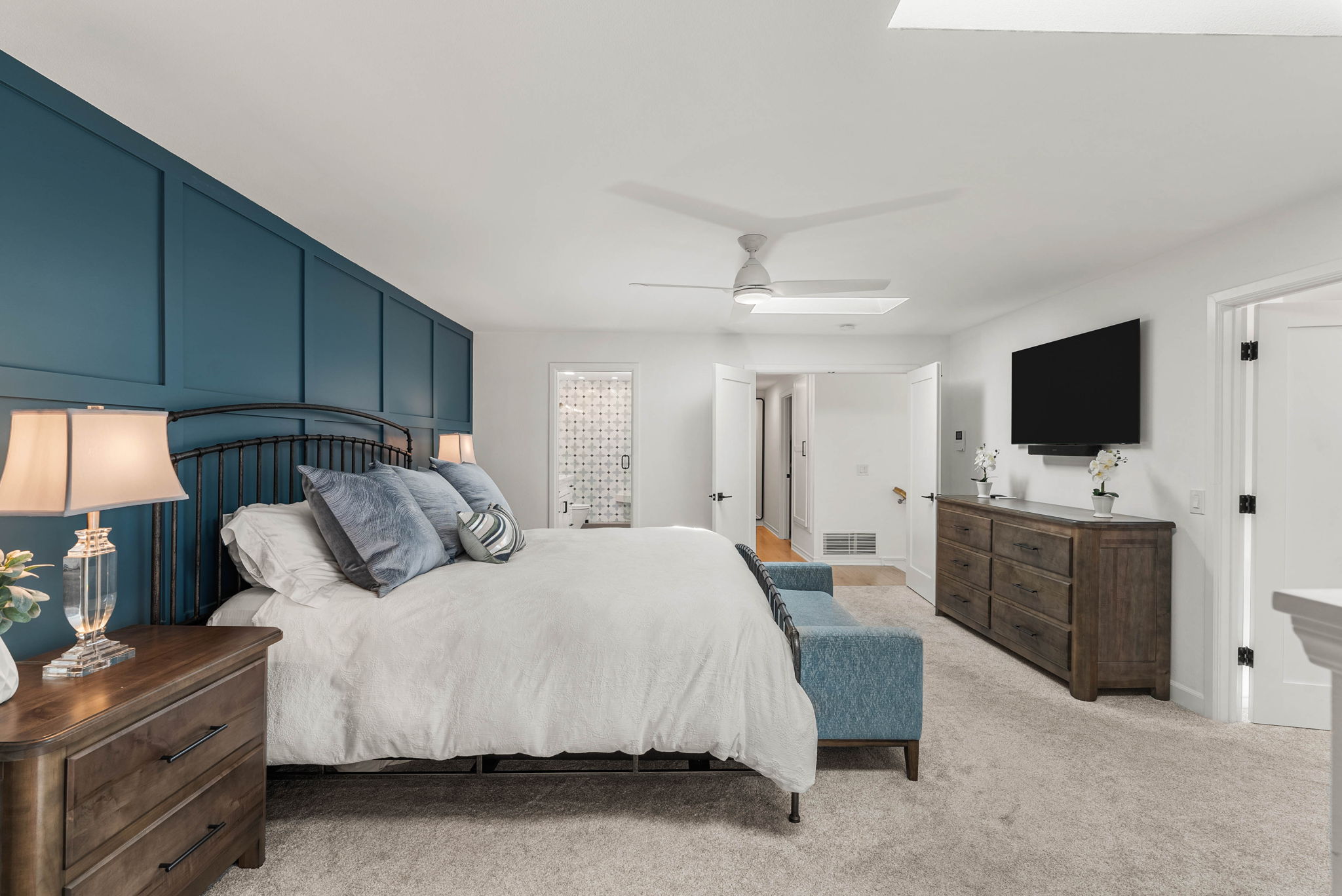 This spacious and inviting primary bedroom features a striking blue accent wall with decorative molding behind a classic metal bed frame. The room is well-appointed with matching wooden dressers, a cushioned bench at the foot of the bed, and neutral carpeting, creating a sophisticated and cozy atmosphere. Double doors lead to an adjoining hallway and a bathroom, adding functional flow and appeal to the suite.