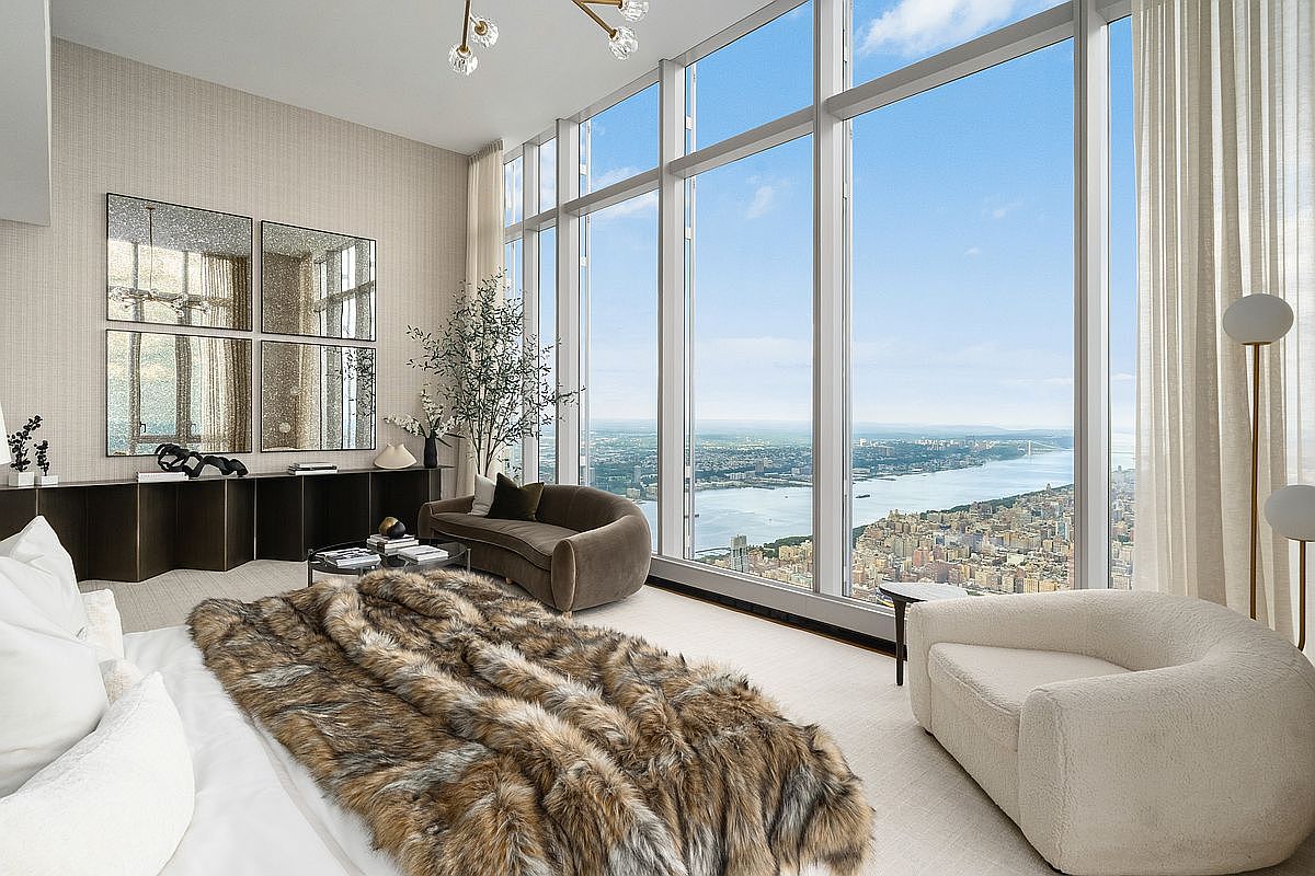 This luxurious primary bedroom features floor-to-ceiling windows that offer a breathtaking, panoramic view of the city skyline and river. The room is elegantly furnished with a plush, textured faux-fur throw on the bed, a curved velvet sofa, and a modern cream-colored armchair, all set against a neutral, sophisticated color palette. The space is accented by a sleek dark console table topped with decorative mirrors, creating an atmosphere of high-end, contemporary urban living.