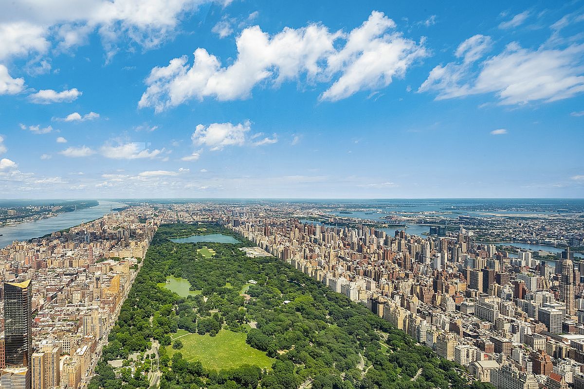 This breathtaking high-altitude aerial view captures the iconic expanse of Central Park in New York City, flanked by the dense, towering skyscrapers of Manhattan. The perspective highlights the stark contrast between the lush, sprawling greenery of the park and the intricate urban grid of the surrounding city, with the Hudson River visible in the distance. It offers a cinematic, panoramic sense of scale and prestige, emphasizing the prime location and architectural density of the area.