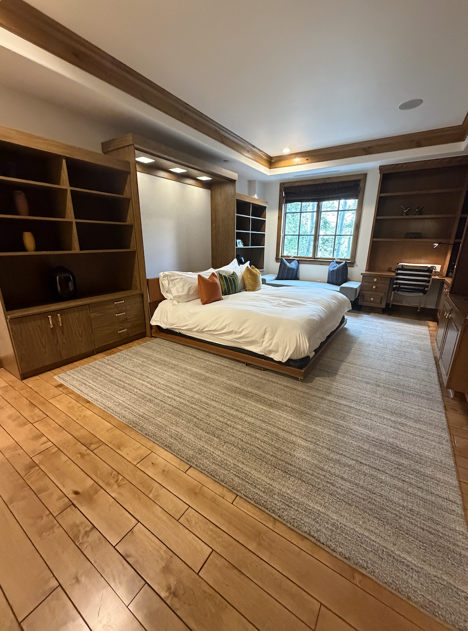 This spacious primary bedroom features custom built-in wooden cabinetry and a central bed flanked by shelving, creating a sophisticated and functional design. A large area rug warms the hardwood flooring, while a cozy window seat and a dedicated desk area provide relaxing and productive spaces. The warm wood tones and balanced layout suggest a high-end, comfortable living environment.
