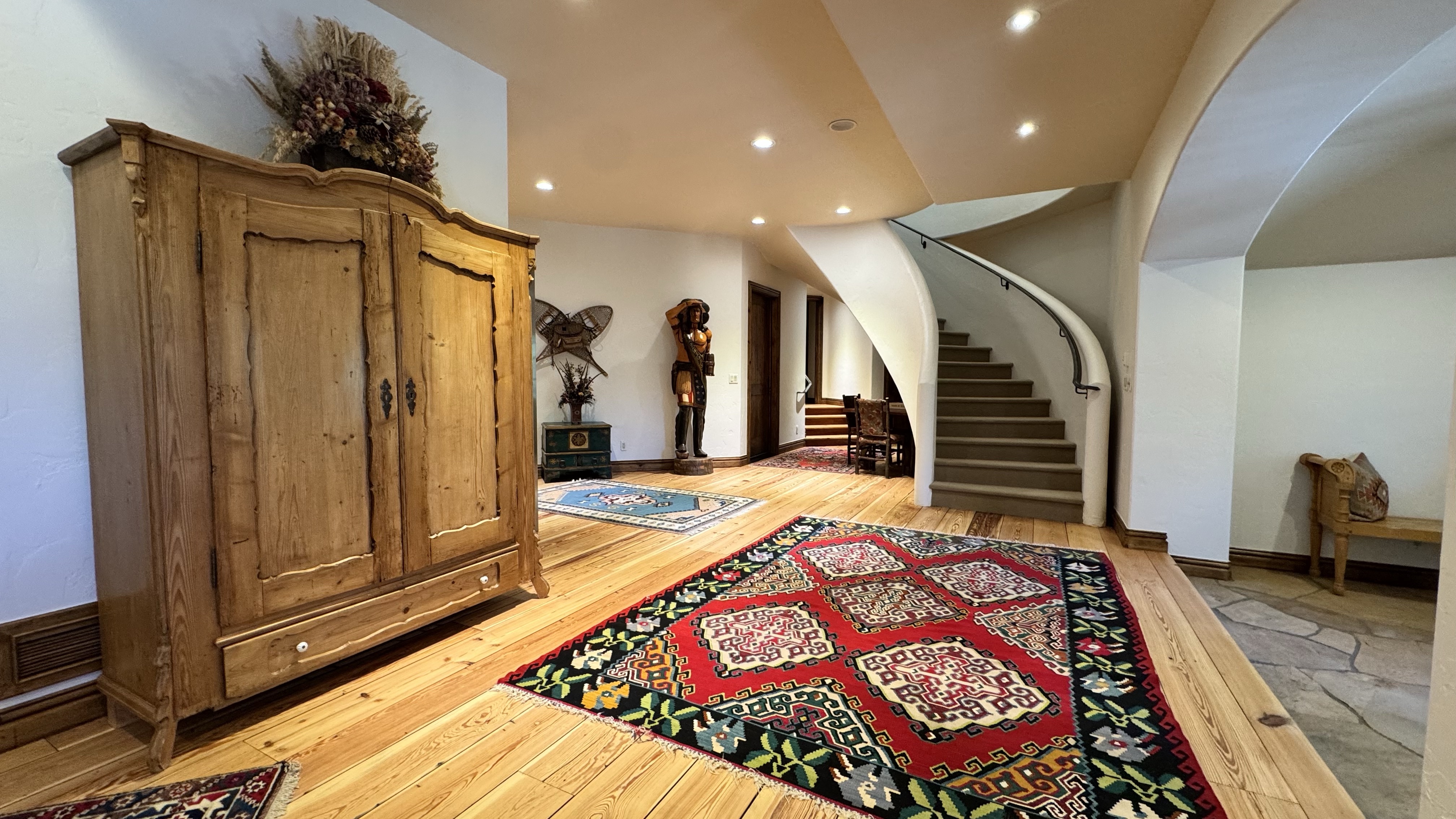 This spacious entryway features a grand, curved staircase with beige carpeting and a sleek railing, serving as the focal point of the area. The room is decorated with warm wood flooring, an antique armoire, and vibrant, patterned area rugs that add color and character to the unique, southwestern-inspired space. The interplay of natural lighting, recessed ceiling lights, and architectural arches creates an inviting and high-end atmosphere.