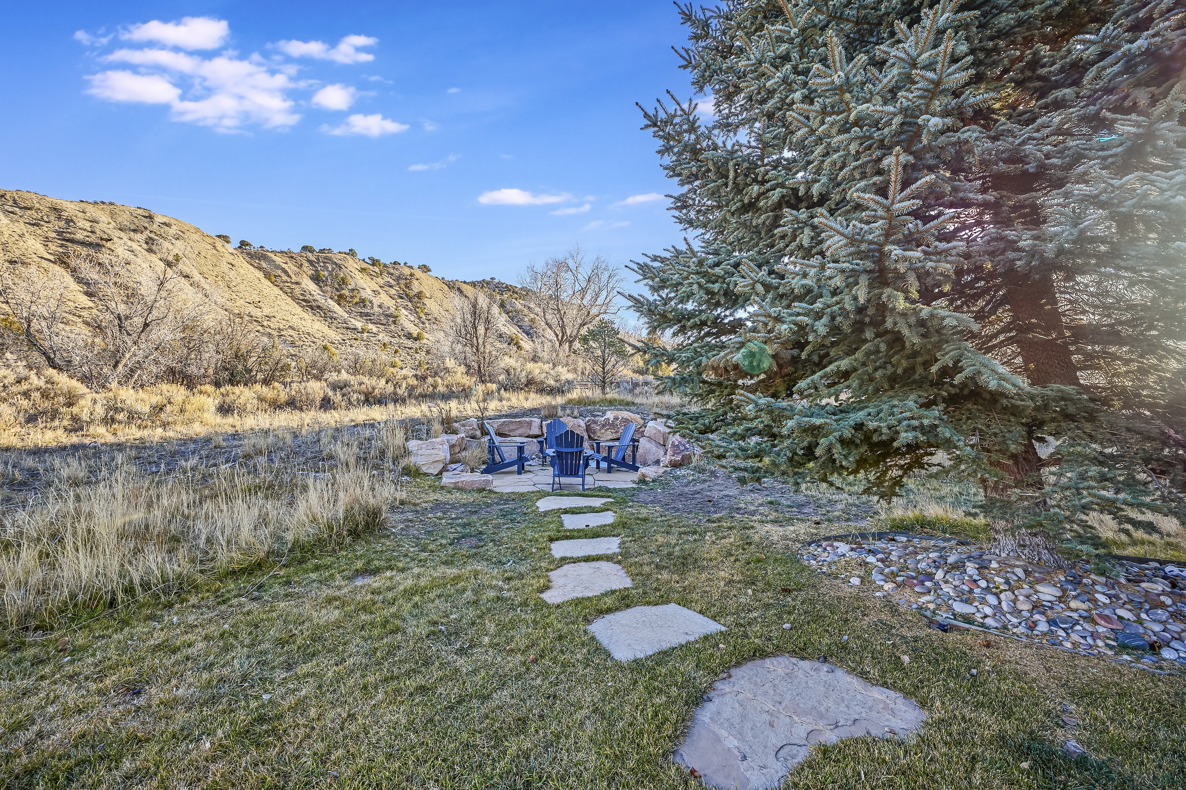 This image showcases a well-maintained yard or garden area, featuring a stone pathway leading to an outdoor seating arrangement. Mature trees provide shade and enhance the landscape. The setting, likely adjacent to a natural, rugged terrain, suggests a peaceful and private outdoor experience.