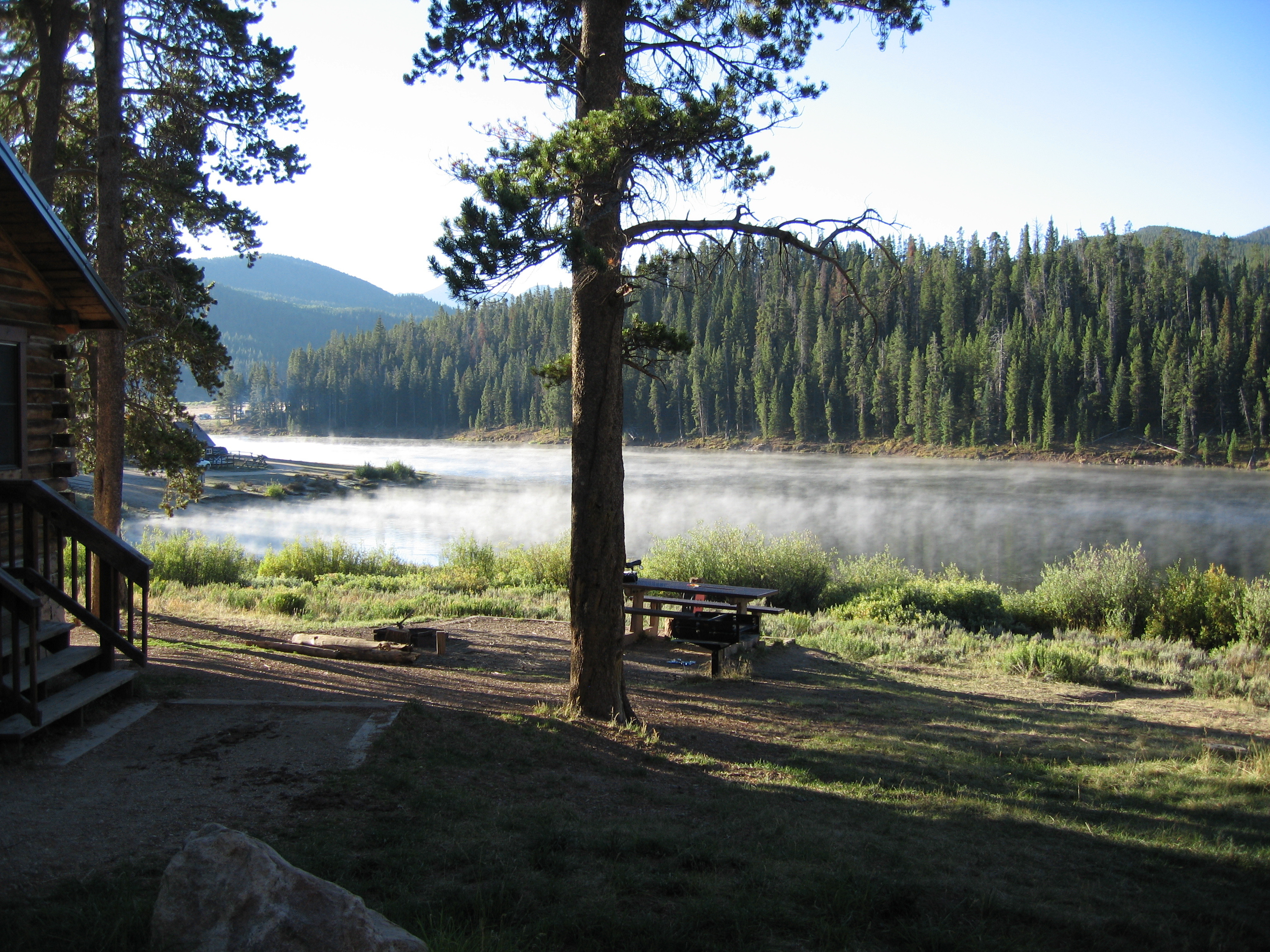This exterior shot showcases a serene yard or garden area next to a rustic cabin. Calm water is visible in the background with light fog rising. A picnic table is situated near a tall tree, suggesting a relaxing outdoor space for gatherings.