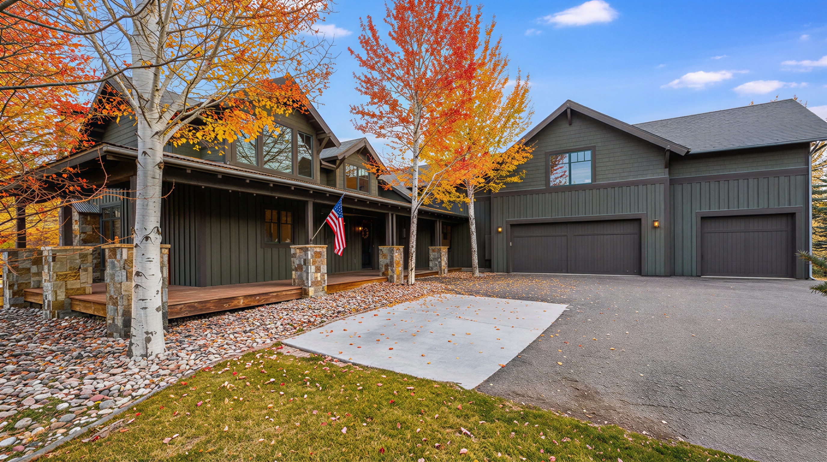 The image showcases the front exterior of a house in the autumn season. The house features a wooden porch with stone pillars, an American flag, and a multi-car garage. The surrounding landscape includes colorful fall foliage, adding to the property's curb appeal.