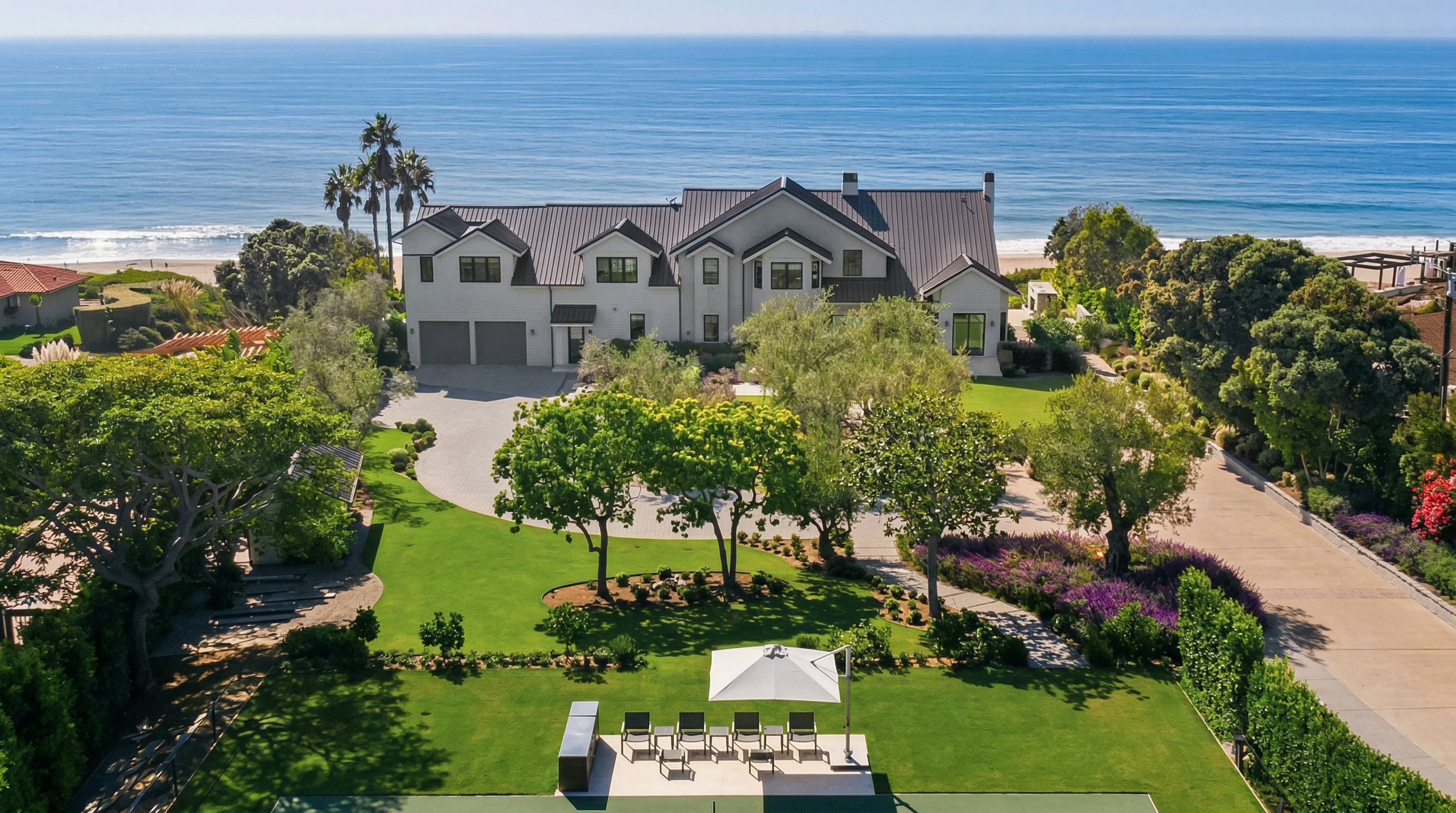 This stunning aerial view captures a large, modern coastal estate set against a vast ocean backdrop. The property features a manicured green lawn, mature trees, a private court, and a spacious circular driveway leading to a multi-car garage. With its elegant architecture and prime beachfront location, this home offers a blend of luxury and tranquility.