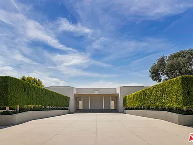 This image showcases the front exterior of a modern residence. The property features a symmetrical design with manicured hedges flanking a paved entryway leading to a covered entrance. The house has a minimalist aesthetic with clean lines and a neutral color palette, creating an inviting yet sophisticated curb appeal.
