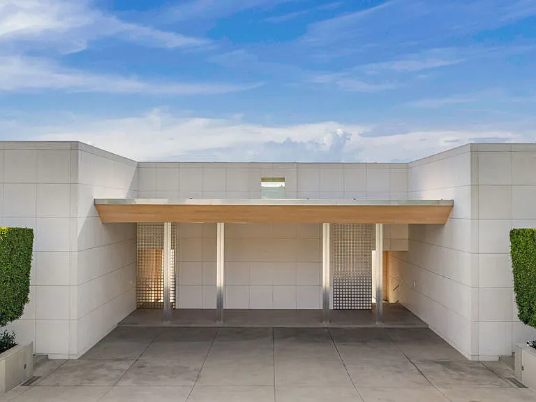 This image showcases the entryway of a modern building with minimalist design. The entrance features a geometric structure composed of white panels, complemented by a light wood overhang and square columns. Green topiaries in rectangular planters flank the entrance, adding a subtle touch of nature to the contemporary architectural style.