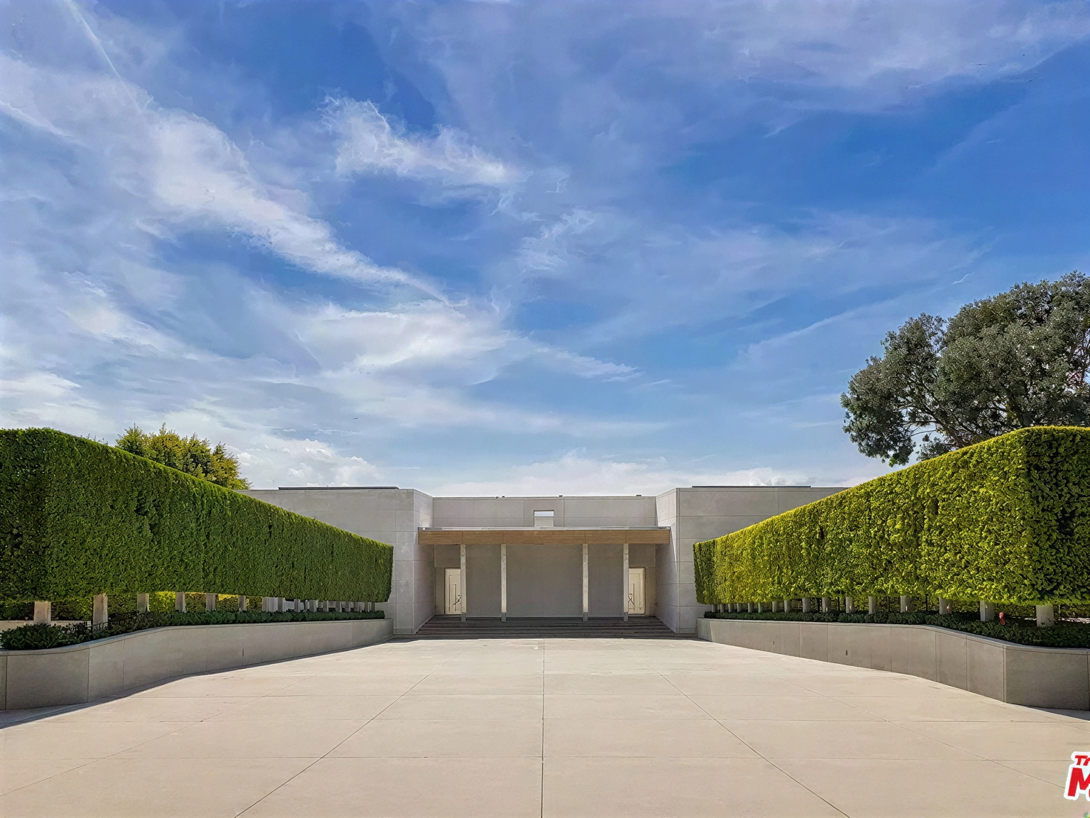The image showcases the front exterior of a modern home with a symmetrical design. A paved driveway leads to a minimalist facade featuring light-colored walls, geometric hedges, and a covered entryway with columns. The architecture emphasizes clean lines and a sense of serene sophistication.