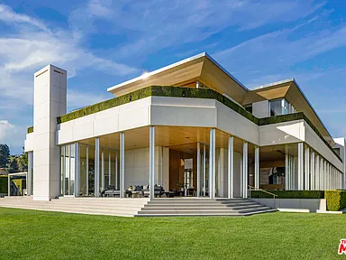 This image showcases the rear exterior of a modern luxury home. The architectural design emphasizes clean lines, with large glass panels providing extensive views and natural light. A manicured lawn and elevated patio with seating areas enhance the outdoor living space.