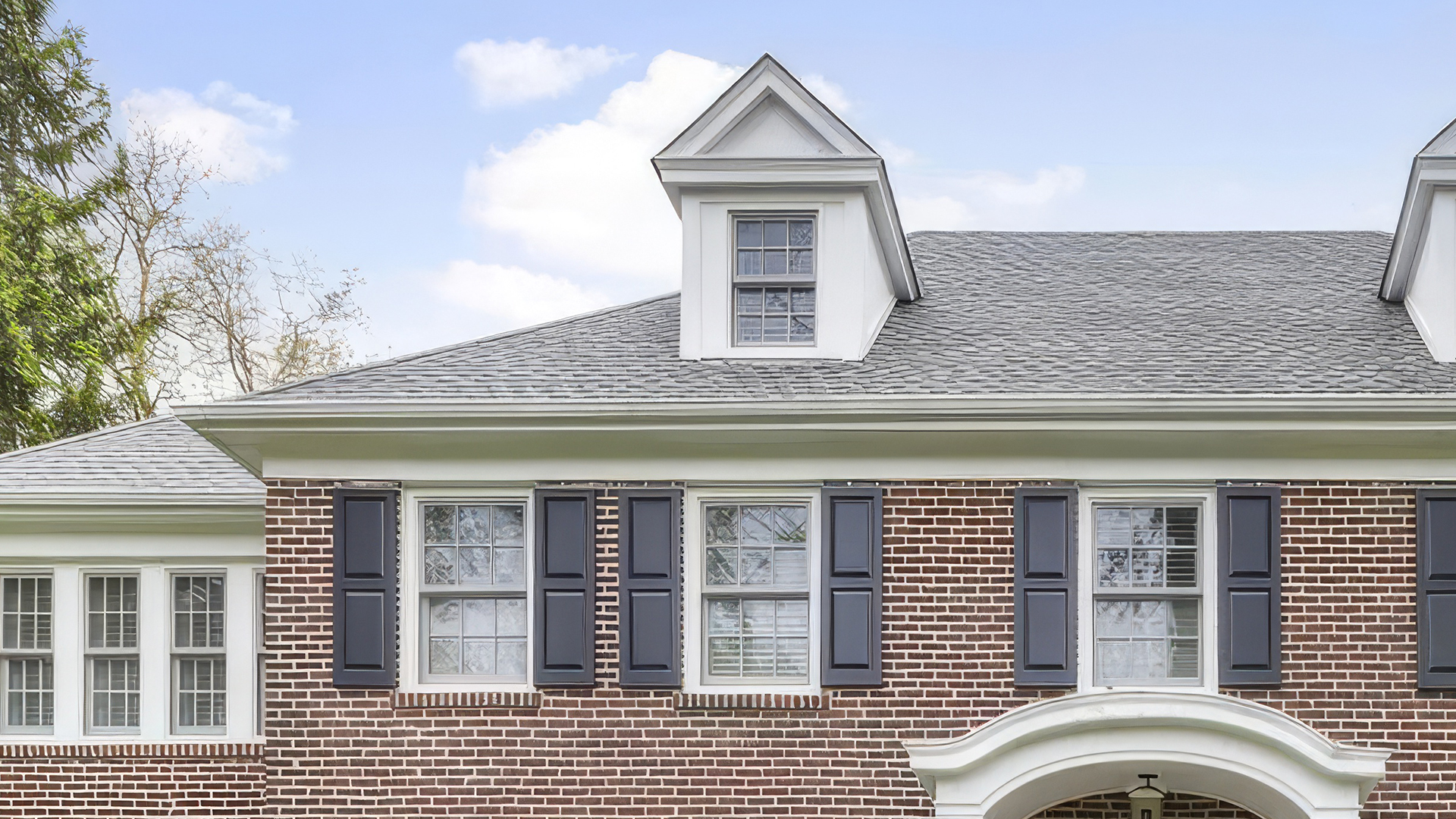 This image showcases the front exterior of a brick house with dark shutters and a gray roof. The architecture includes dormer windows and a covered entryway, suggesting a traditional style. The yard is partially visible and features greenery.