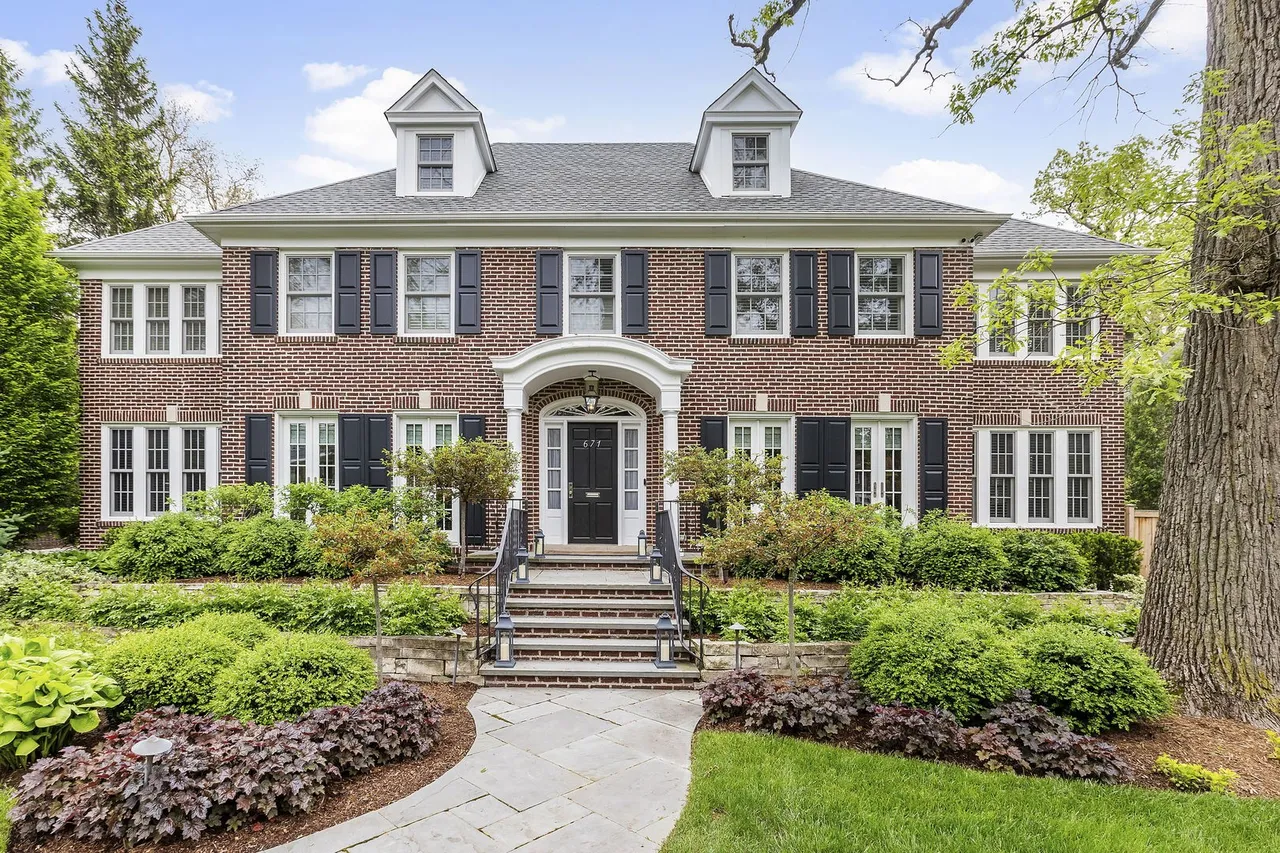 This is an appealing front view of a stately brick home. The symmetrical facade features black shutters, white-framed windows, and dormers on the roof. Lush landscaping adds to the curb appeal, creating an inviting and well-maintained presence.
