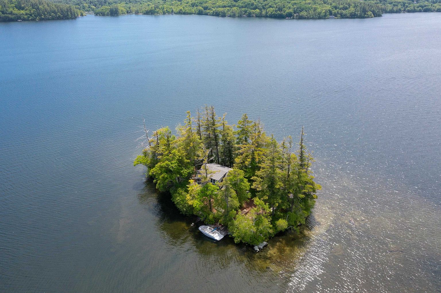 This high-angle aerial view captures a secluded private island nestled in a large, calm lake. The island is densely wooded with mature trees surrounding a single residential structure, and a small motorboat is docked at a modest pier along the shoreline. The perspective emphasizes the property's unique isolation and serene natural setting, offering a tranquil retreat from the mainland visible in the background.