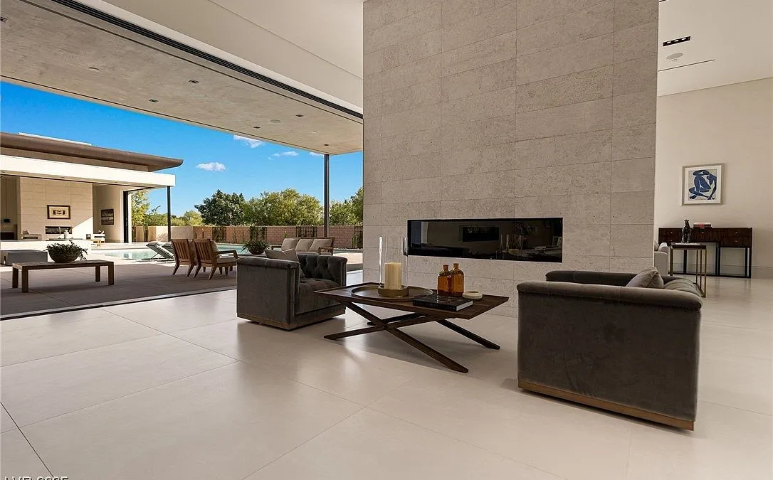 This interior shot showcases a luxurious living room in a modern home. The room features an open floorplan that leads to an outdoor patio, with access to a pool area enhancing the indoor-outdoor flow. The color palette is neutral, with contemporary furnishings complemented by a sleek fireplace feature, making the space feel sophisticated and inviting.