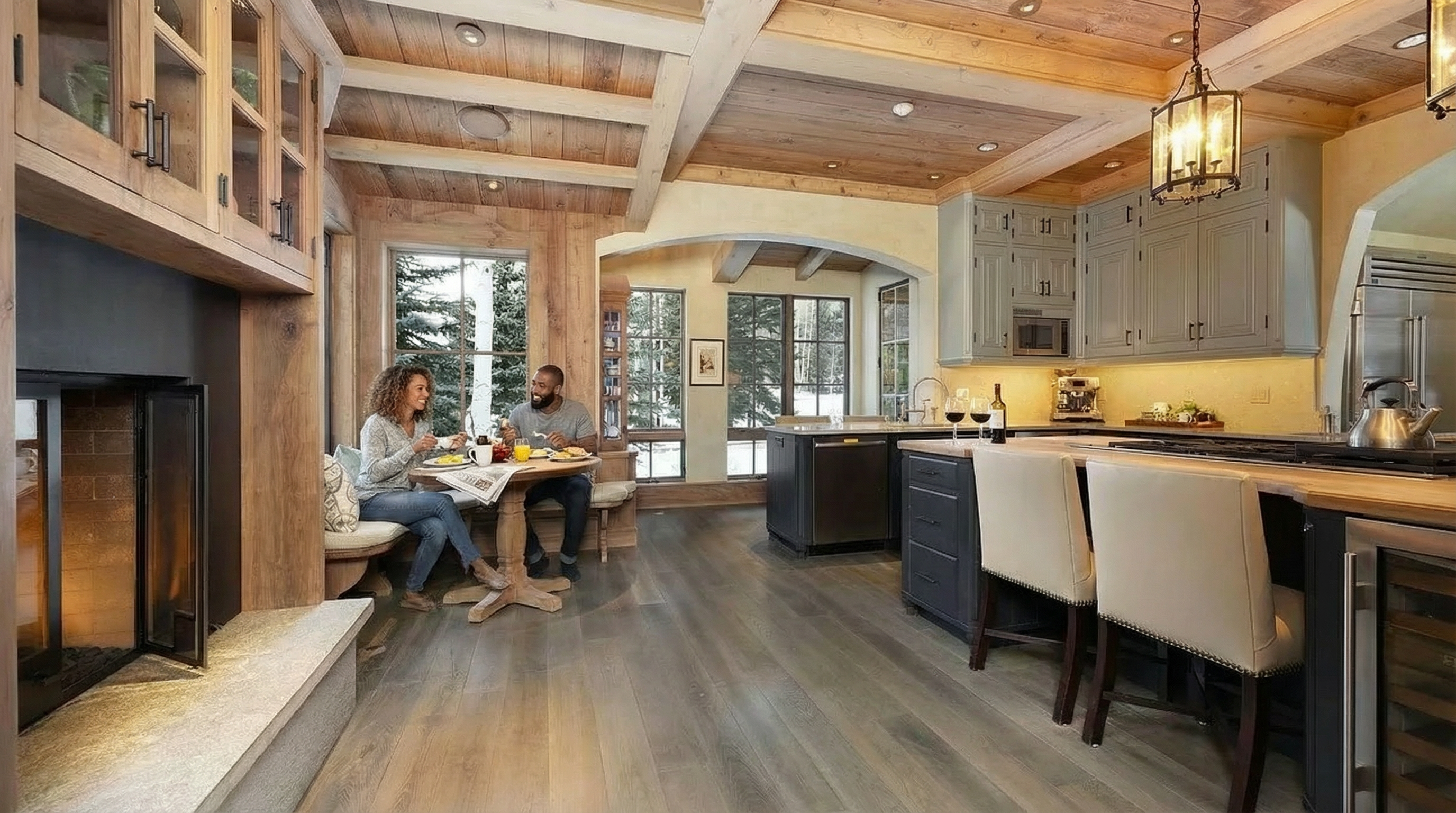 This is an inviting kitchen with rustic and modern touches. It features wood beamed ceilings, gray cabinetry, and an island with seating. It also contains a table where a couple is eating, adding to the cozy atmosphere of the space.