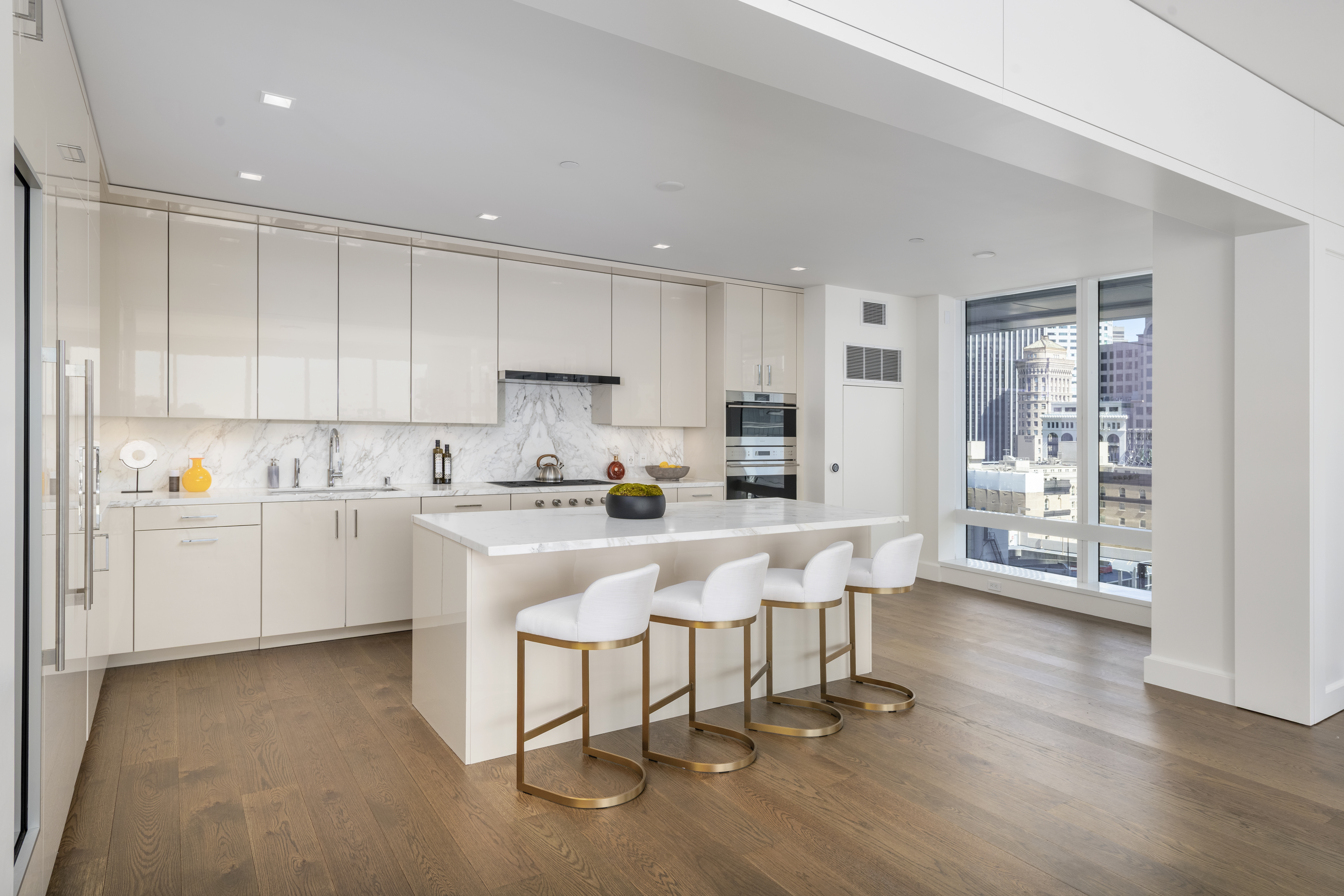 This sleek, contemporary kitchen features high-gloss cream cabinetry paired with elegant marble countertops and a matching backsplash. A spacious central island provides seating for four with stylish white and brass bar stools, while large windows flood the area with natural light and showcase expansive city views. The space offers a clean, sophisticated aesthetic complemented by rich hardwood flooring throughout.