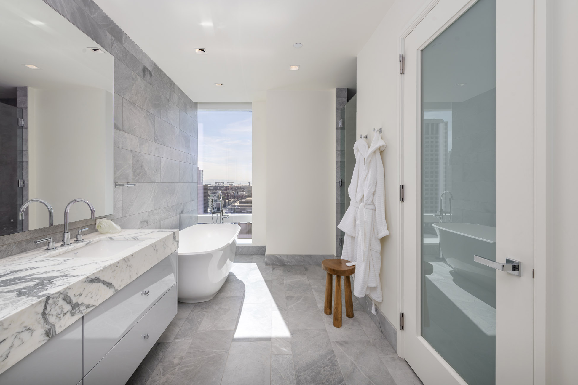 This modern, luxury primary bathroom features a sleek marble vanity, a deep freestanding soaking tub, and floor-to-ceiling tile work in a sophisticated gray palette. Large windows provide ample natural light and expansive city views, while a frosted glass door adds architectural detail. The space is uncluttered and contemporary, emphasizing high-end finishes and a clean, serene environment.