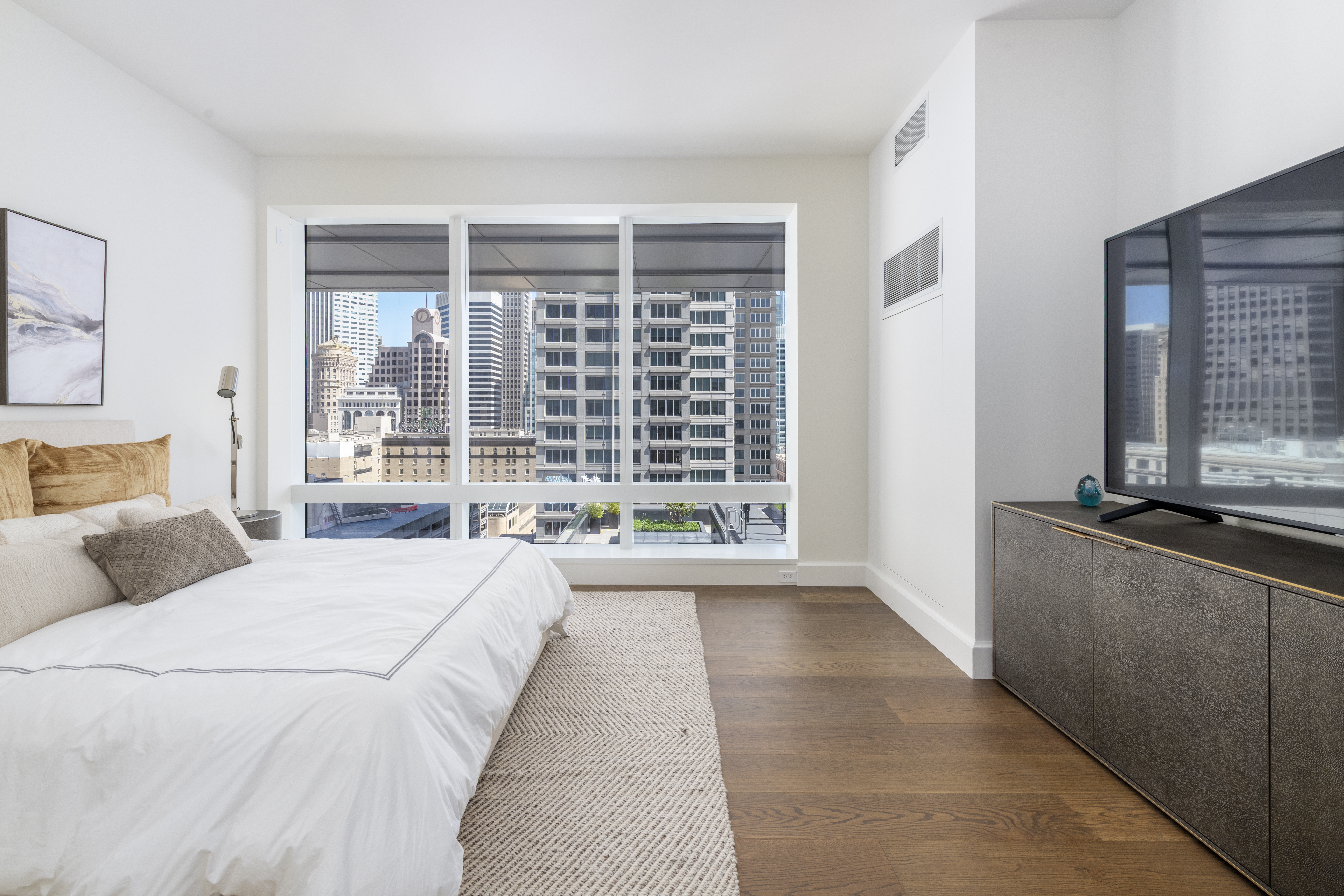 This modern primary bedroom features floor-to-ceiling windows that provide striking urban city views, filling the minimalist space with natural light. The room is appointed with a plush, textured bed featuring a soft neutral color palette, a sleek media console, and rich dark wood flooring. The overall aesthetic is contemporary, sophisticated, and serene, perfect for an upscale metropolitan high-rise residence.