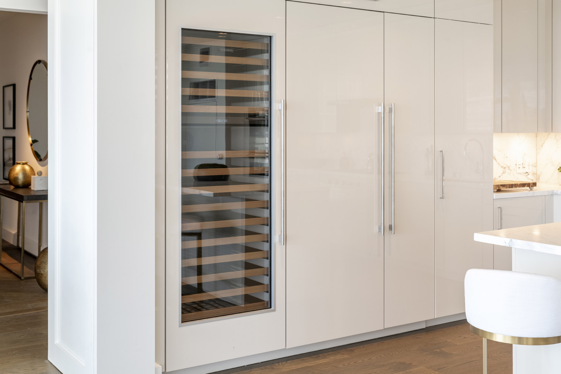 This kitchen features a sleek, floor-to-ceiling cabinetry wall with a built-in, glass-front Sub-Zero wine refrigerator. The design is modern and high-end, characterized by minimalist white panels, long silver hardware, and integrated appliances that create a seamless look. A glimpse of the adjacent dining area and a marble-topped kitchen island indicate a luxurious, open-concept living space.