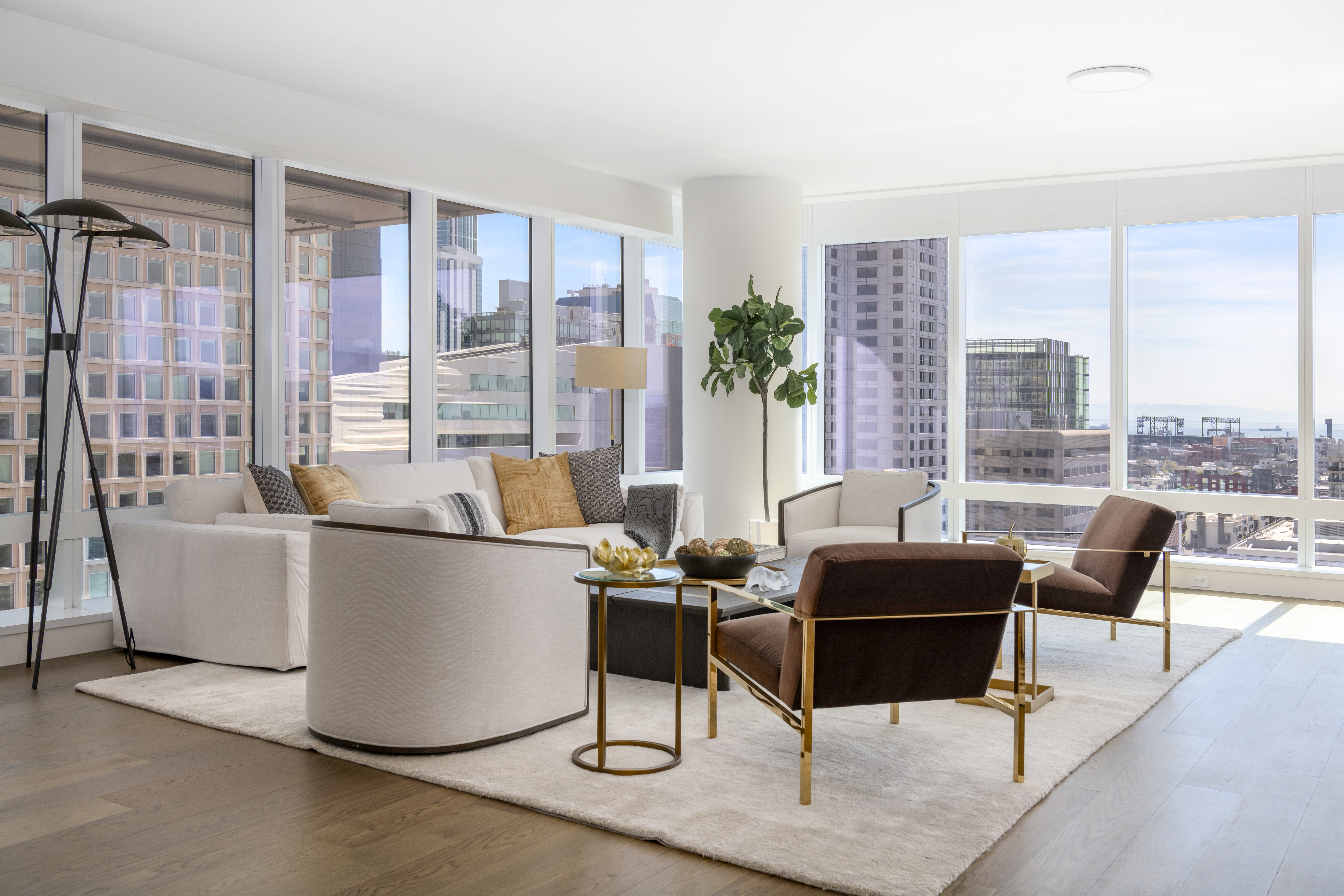 This modern, light-filled living room features floor-to-ceiling windows that provide expansive city views and ample natural light. The room is styled with a white sectional, elegant brown accent chairs with gold frames, and a minimalist coffee table, all set upon a neutral area rug. Its sophisticated, high-rise aesthetic is further enhanced by contemporary lighting and an open-concept layout, making it an ideal space for urban living.