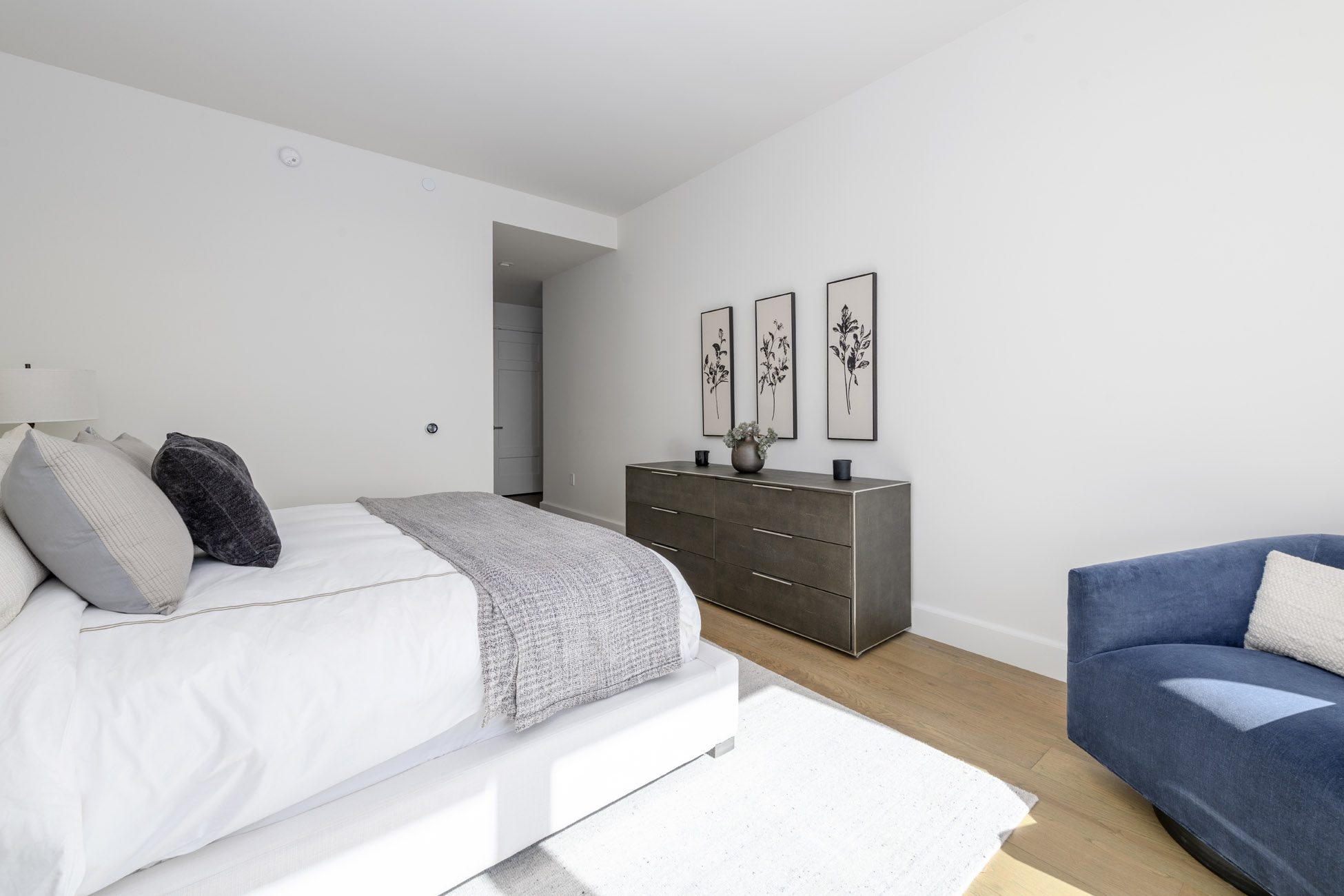 This primary bedroom features a modern, minimalist design with a clean white palette and soft gray and blue accents. The room includes a bed with layered bedding, a metallic six-drawer dresser adorned with floral wall art, and a plush blue armchair, all set against high ceilings and hardwood flooring.