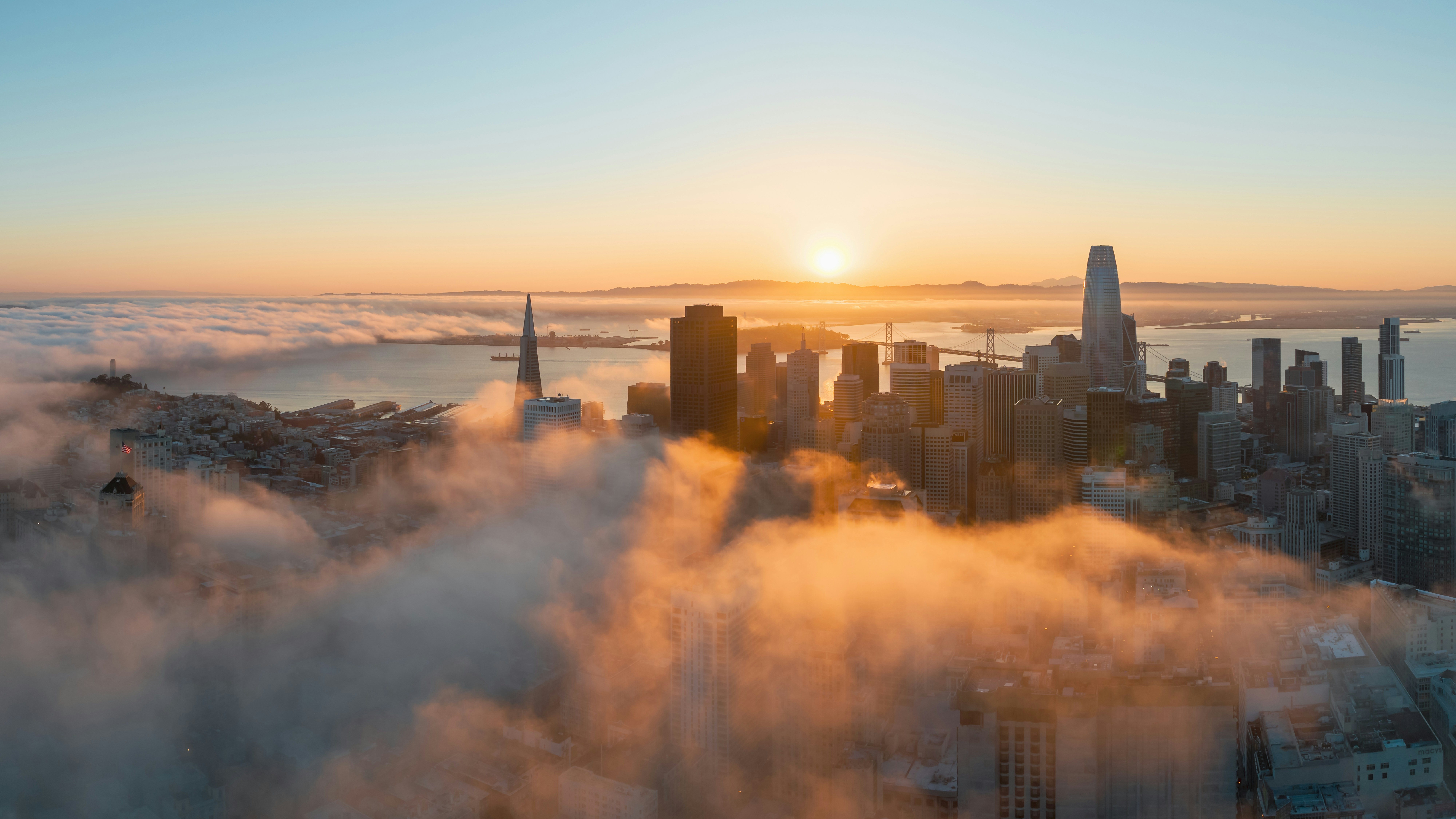 This stunning wide-angle aerial shot captures the iconic San Francisco skyline at sunrise, partially shrouded by a thick blanket of rolling fog. The golden light illuminates the skyscrapers, including the Salesforce Tower, and creates a dramatic contrast with the coastal mist over the bay and the Bay Bridge in the distance. This image highlights the prestigious urban setting and unrivaled panoramic city views available from high-rise vantage points in the area.