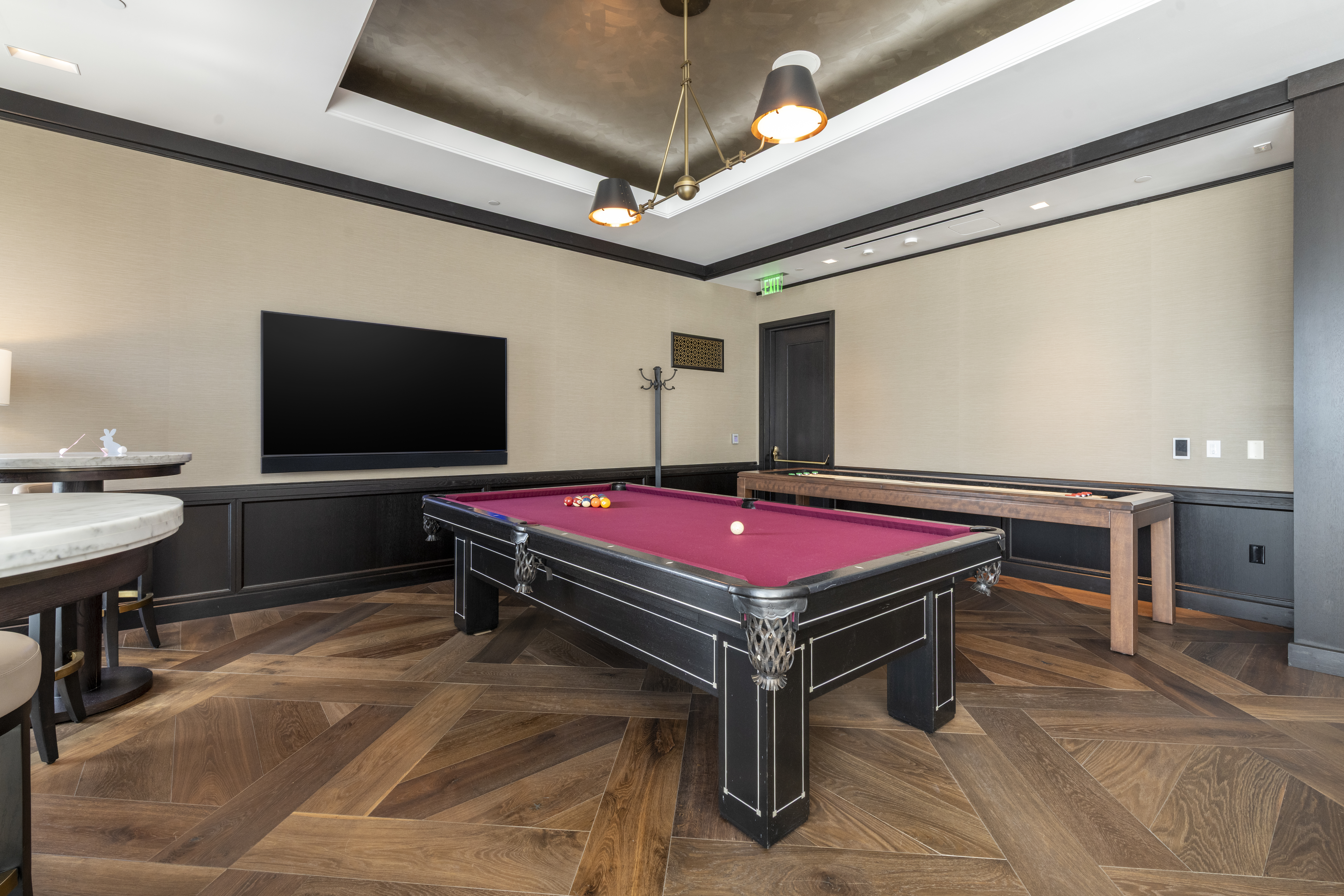 This sophisticated gaming lounge, likely part of an upscale residential amenity space, features a large burgundy felt pool table and a secondary shuffleboard table. The room is designed with high-end finishes, including a multi-tiered tray ceiling with custom lighting, dark wood paneling, and intricate herringbone-patterned hardwood floors. A large flat-screen television and bar seating area complete the luxury recreational setting.