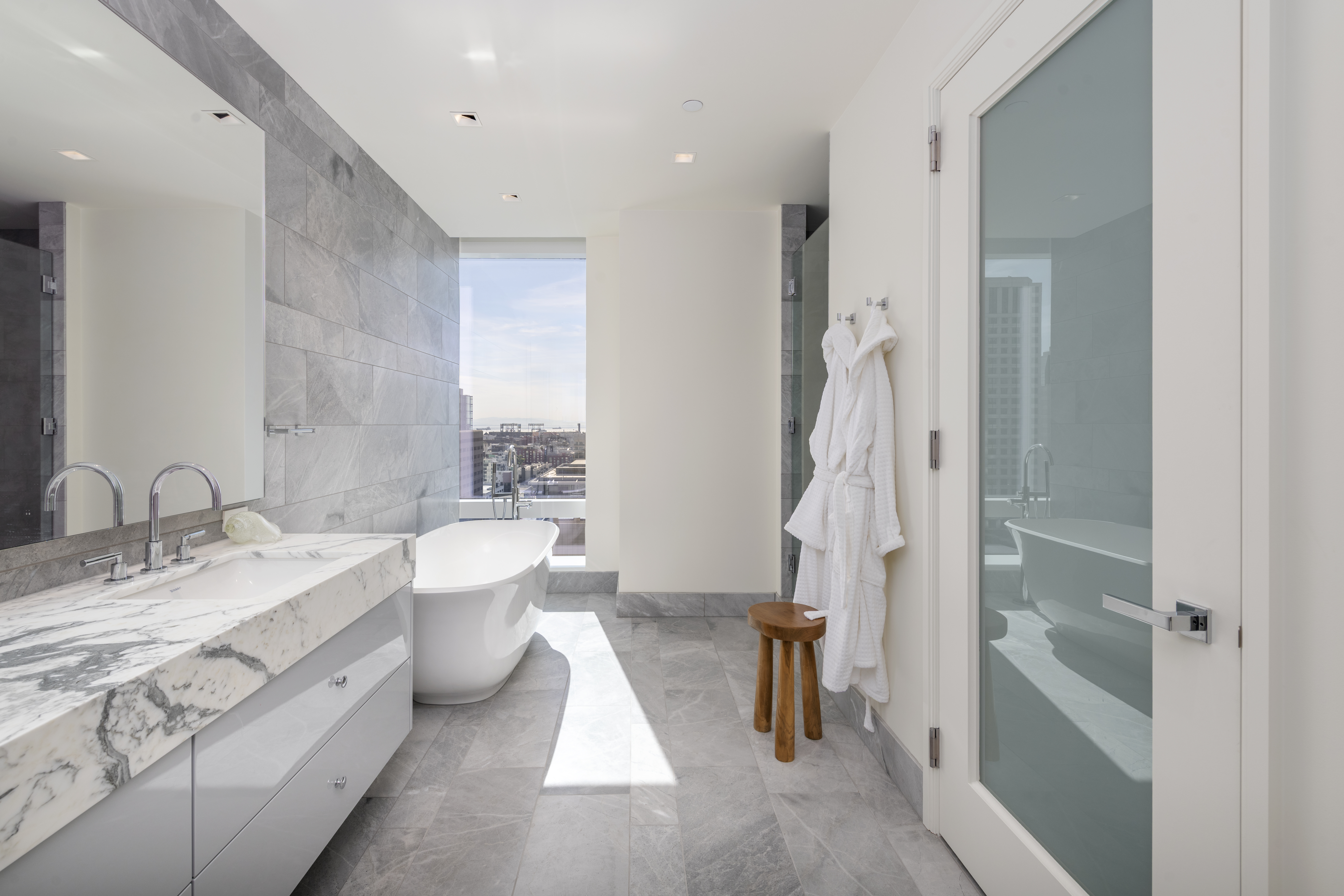 This modern, high-end primary bathroom features a sleek free-standing soaking tub positioned before a floor-to-ceiling window with impressive city views. The space is anchored by a sophisticated vanity with Calacatta marble countertop and polished grey tile flooring, complemented by a minimalist stool and plush white bathrobes. Its clean lines and neutral color palette create an elegant, tranquil spa-like atmosphere.