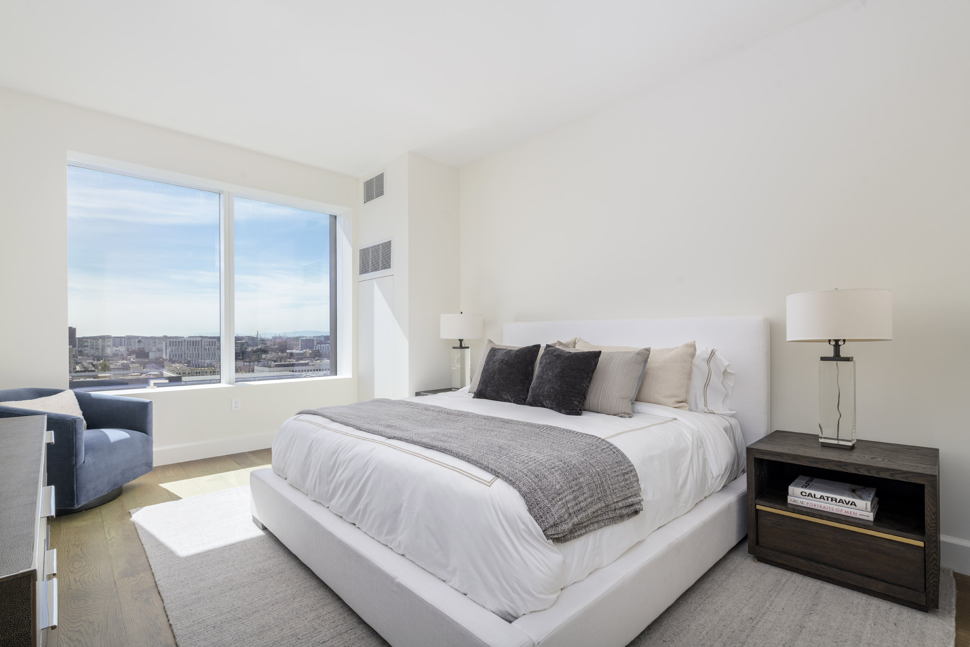 This bright and airy primary bedroom features a crisp, modern aesthetic highlighted by floor-to-ceiling windows that provide expansive urban views. The room is anchored by a king-sized bed with neutral, sophisticated linens and flanked by dark wood nightstands with elegant lamps. A plush blue armchair in the corner adds a touch of color to the otherwise monochromatic, minimalist design.