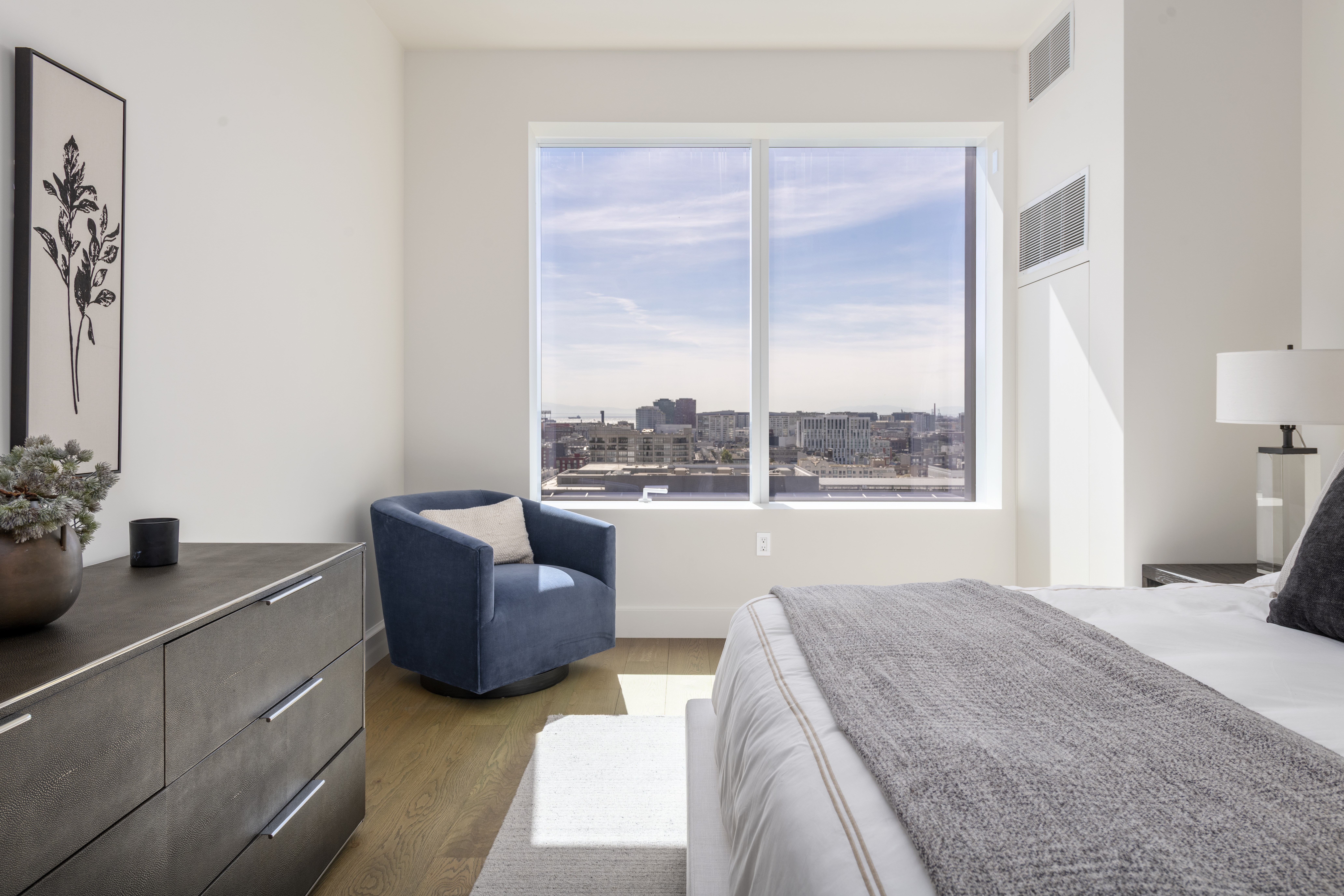 This sleek, modern primary bedroom features a tranquil atmosphere highlighted by floor-to-ceiling windows that provide expansive urban views. The room is appointed with a textured grey dresser, a chic blue swivel accent chair, and a neutral-toned bed with premium linens. The combination of high-end finishes, minimalist decor, and natural light creates a sophisticated and calming living space.