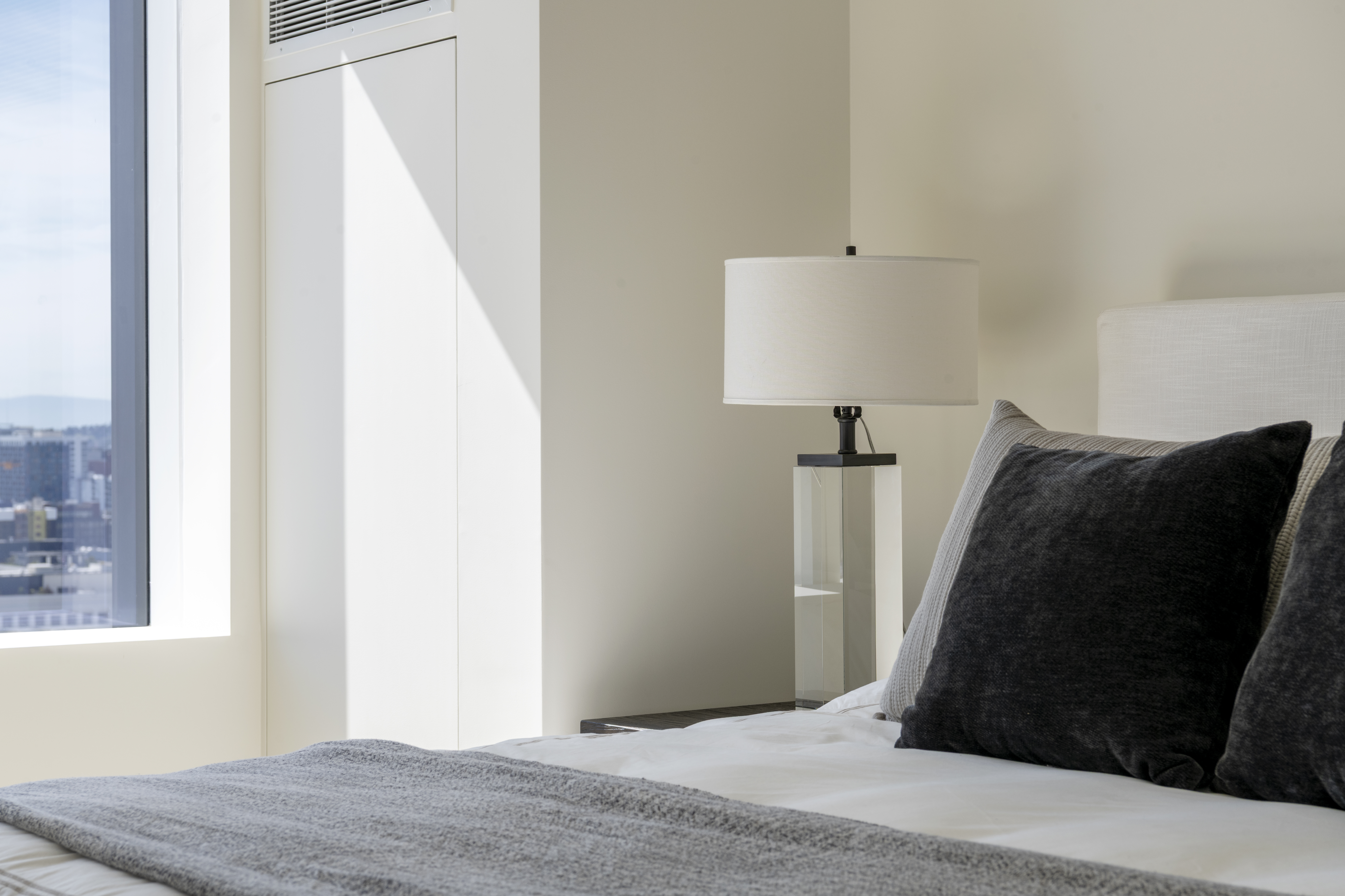 This serene bedroom features a contemporary design highlighted by soft neutral tones and clean lines. A sleek, transparent-base table lamp sits atop a nightstand, while a plush bed with textured accent pillows adds depth and comfort to the room. The space is naturally brightened by a large window that offers an expansive urban cityscape view, creating a sophisticated and tranquil atmosphere.