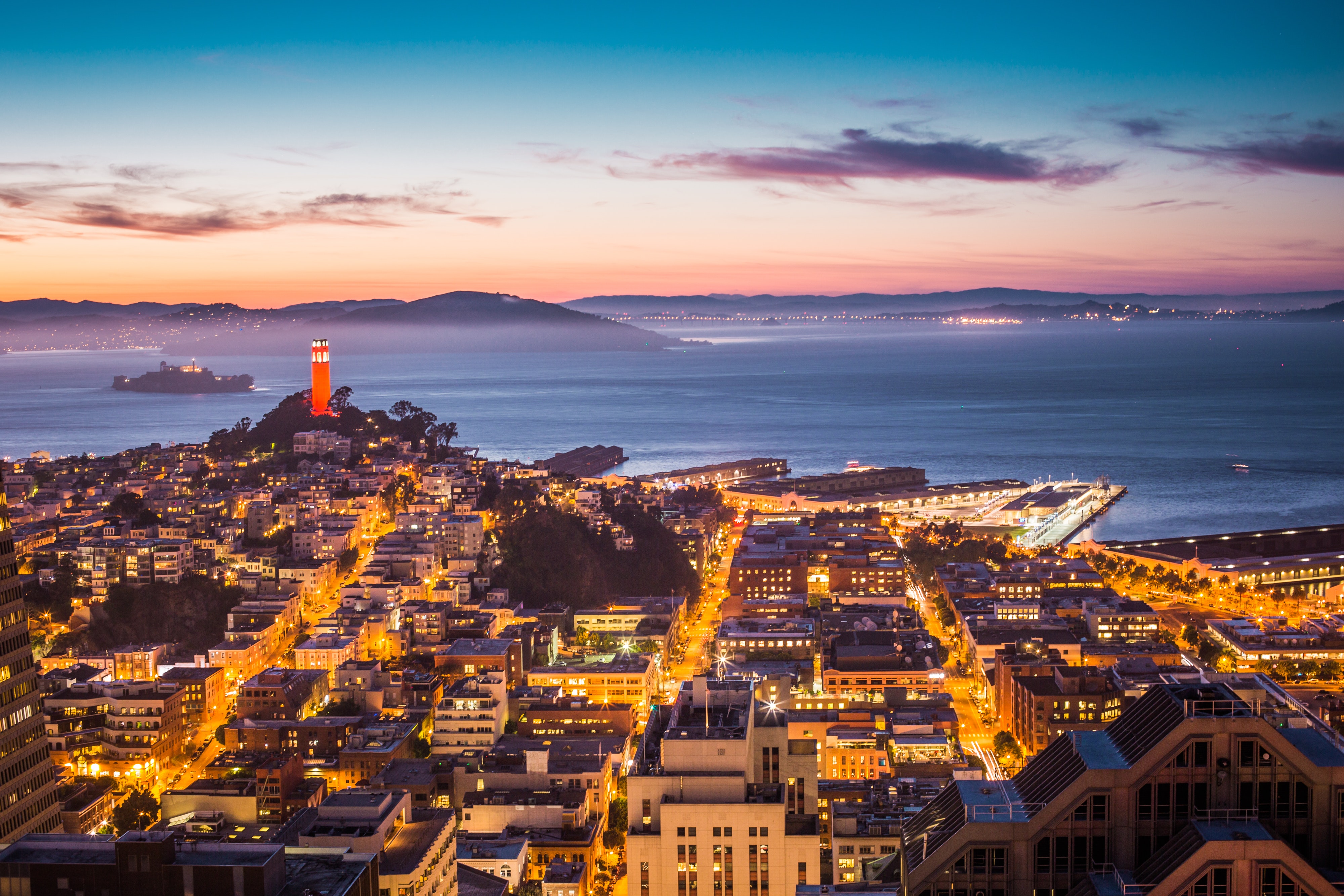 This stunning high-angle aerial view captures a vibrant cityscape at dusk, featuring the iconic Coit Tower prominently illuminated against a colorful twilight sky. The dense urban landscape overlooks the San Francisco Bay, showcasing a blend of historic architecture, busy streets, and active waterfront piers. The composition highlights the city's prime coastal location and offers an expansive perspective of the neighborhood's density and local character.
