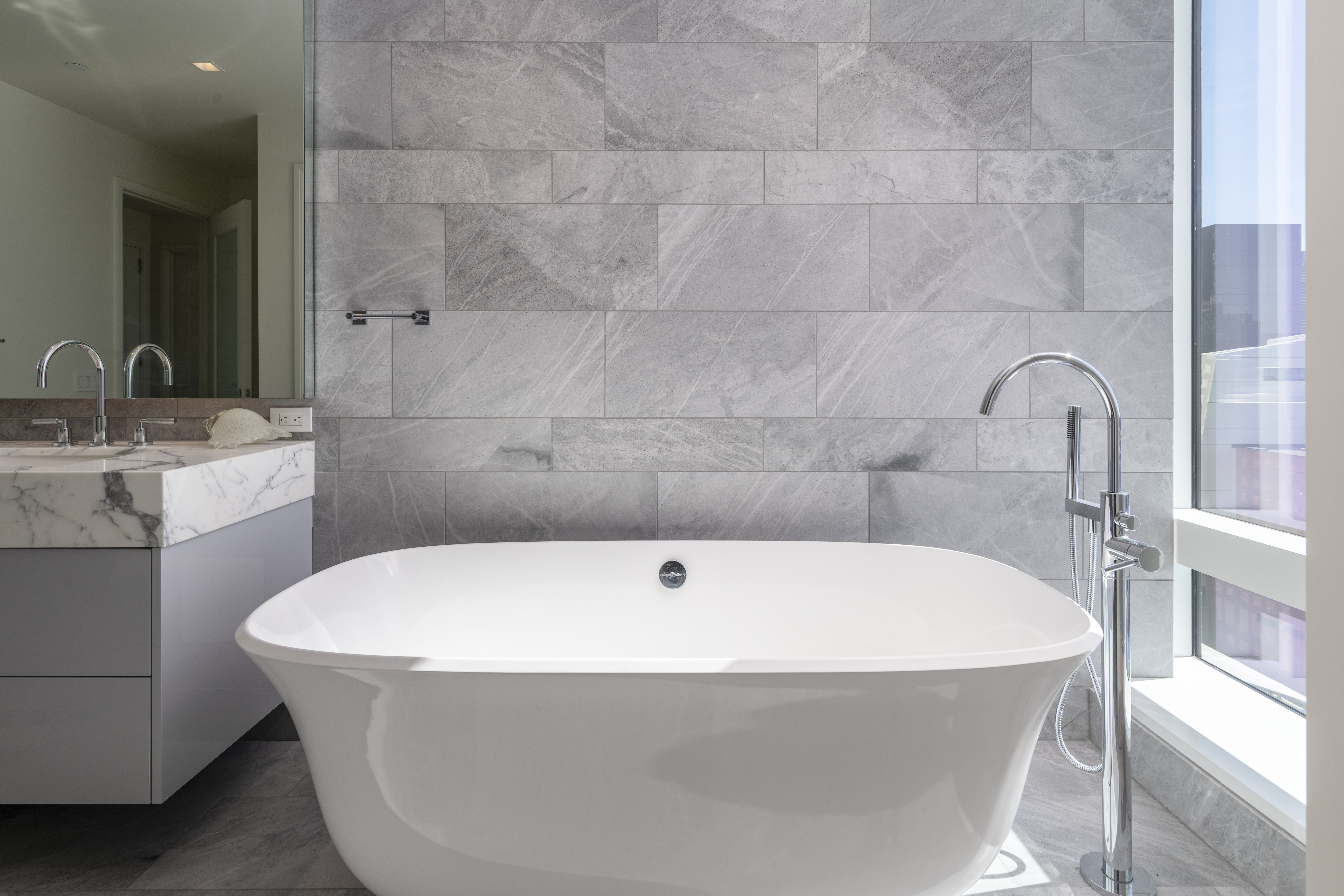 This modern primary bathroom features a sleek freestanding soaking tub paired with a contemporary floor-mounted chrome faucet. The space is accented by a stylish marble-topped floating vanity and elegant gray large-format stone tile walls, creating a minimalist and spa-like atmosphere. Large windows to the side allow for ample natural light, enhancing the bright and luxurious feel of the room.