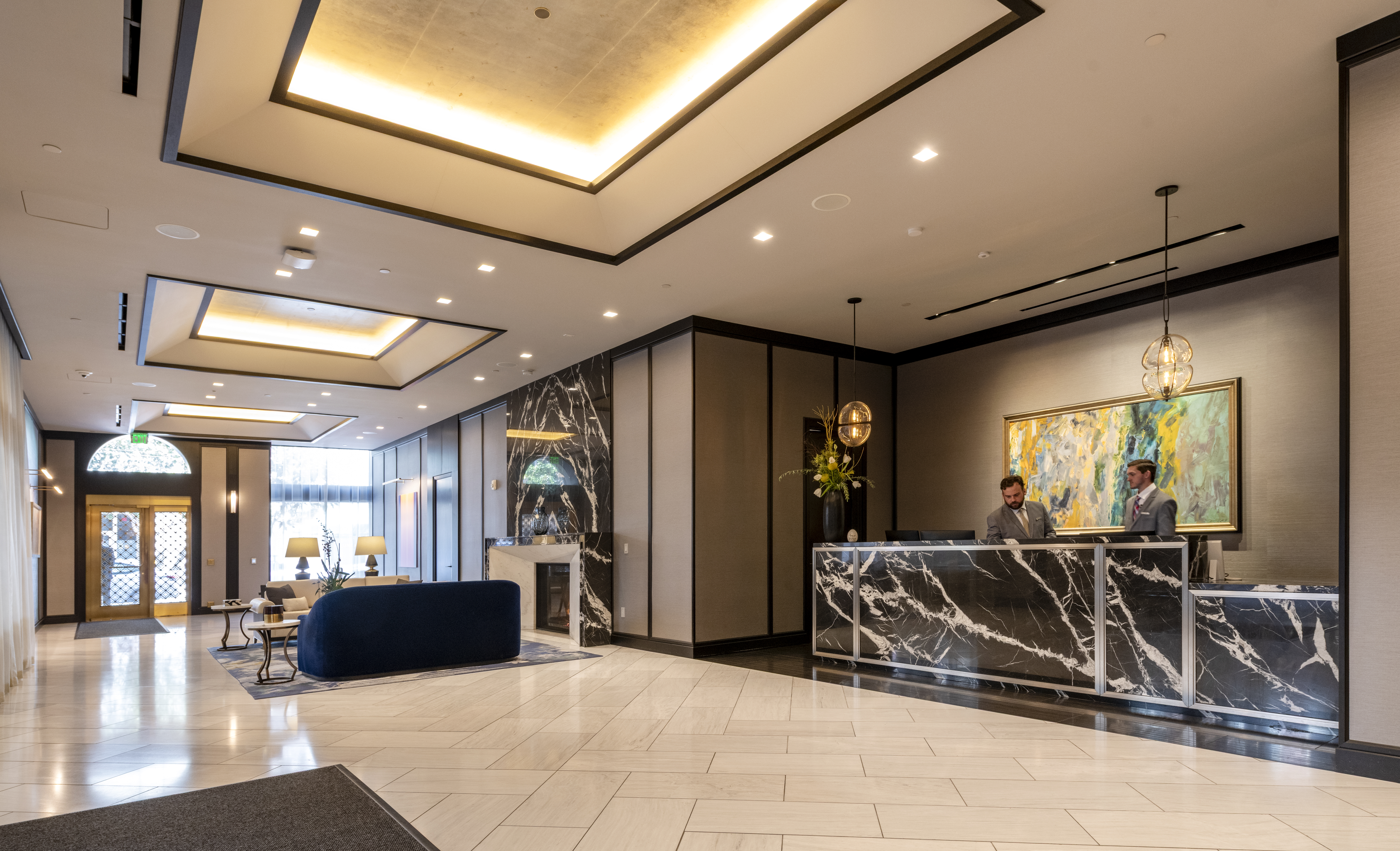 This sophisticated hotel lobby features high ceilings with recessed lighting, a striking black marble reception desk, and polished tile flooring. A cozy seating area with a blue sofa and fireplace creates a welcoming atmosphere, further enhanced by modern artwork and elegant pendant light fixtures. The space is professional, upscale, and conveys a luxury reception experience.