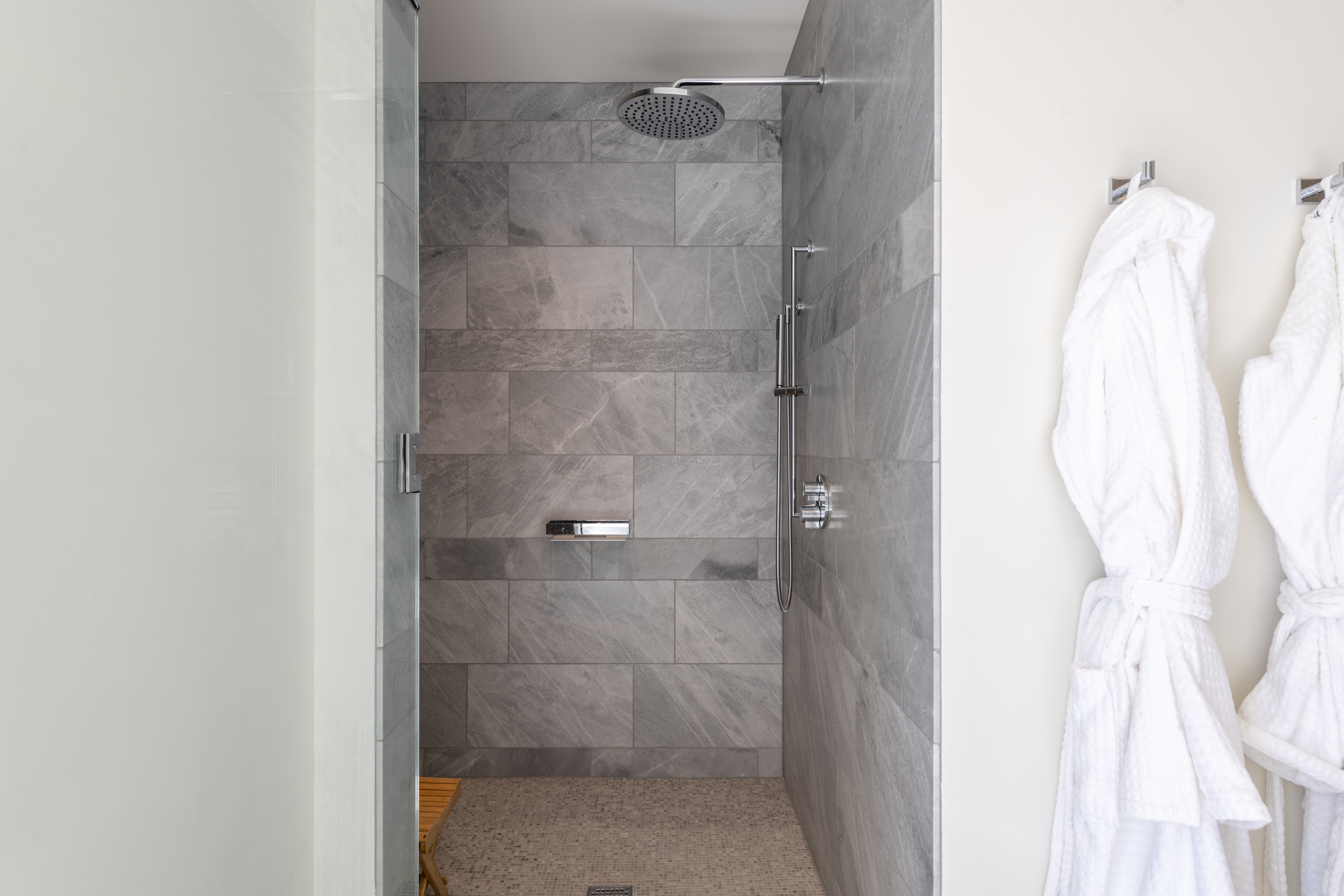 This modern walk-in shower features sophisticated grey stone tiles and a sleek rainfall showerhead paired with a handheld sprayer. A convenient integrated shelf and a corner teak bench enhance functionality, while two plush white bathrobes hanging nearby suggest a luxury, spa-like atmosphere.